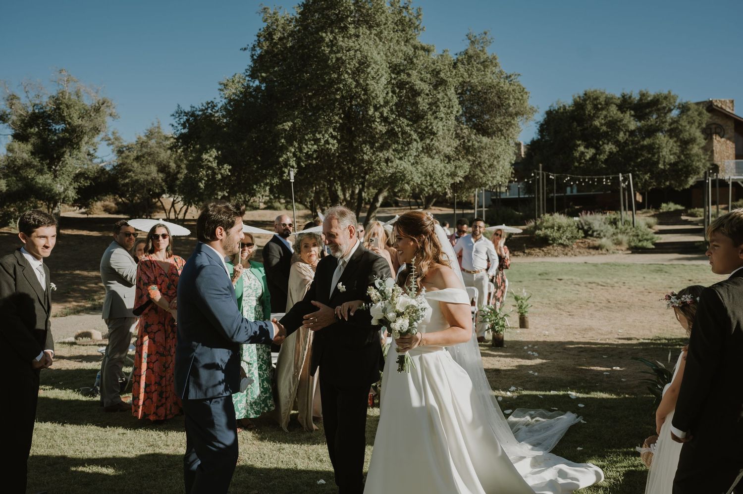 Father walks bride down the aisle at sunny outdoor wedding ceremony with guests watching.