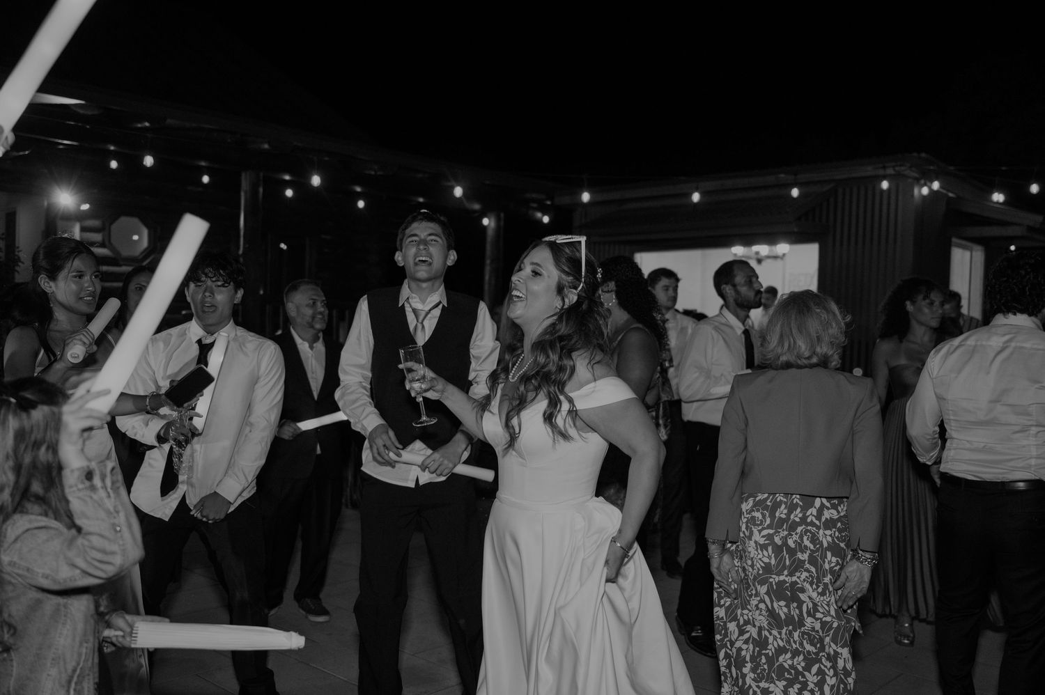 Bride and groom enjoy outdoor wedding reception with guests dancing under string lights at night.