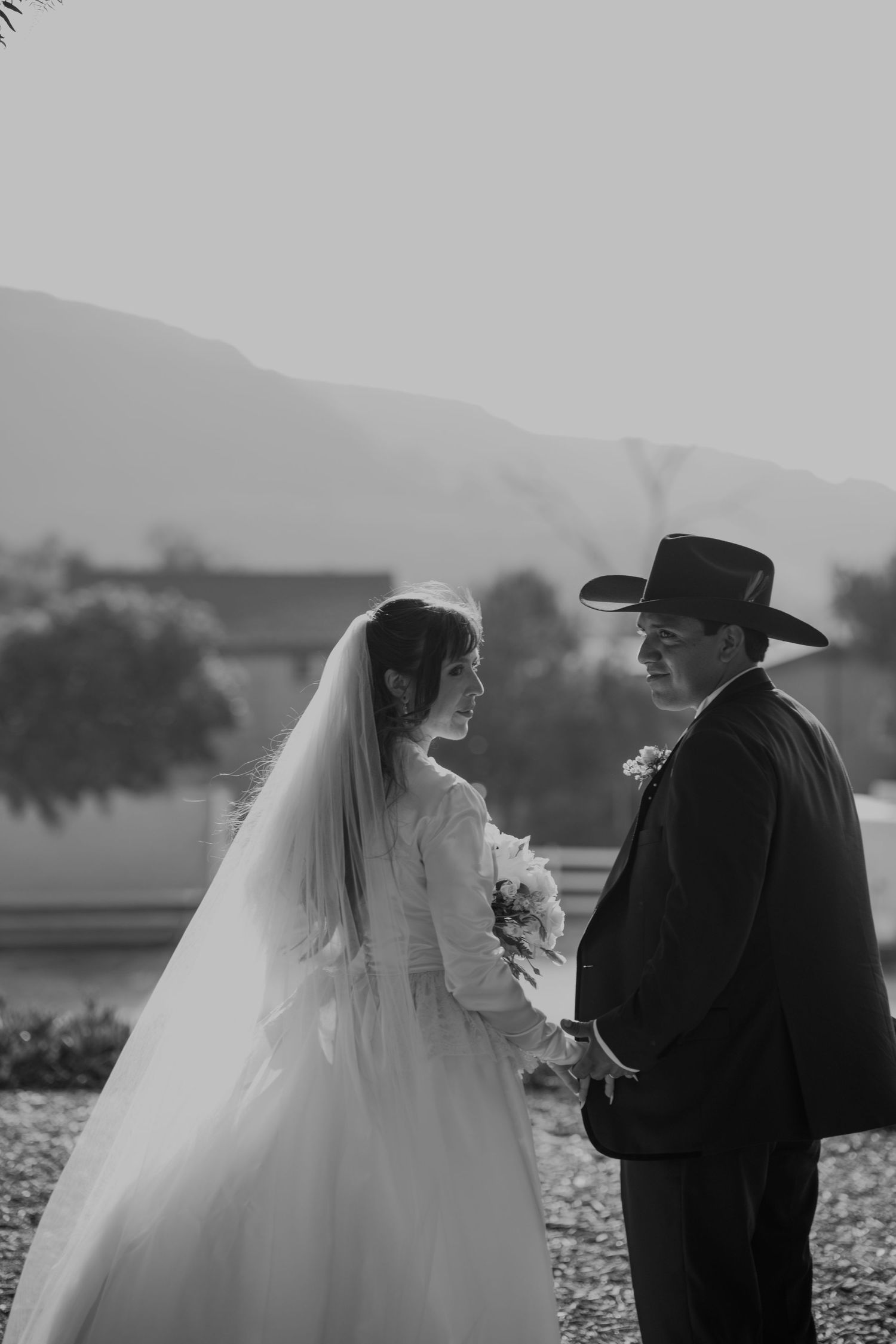 Black and white wedding photo of bride in veil holding bouquet and groom in cowboy hat holding hands outdoors.