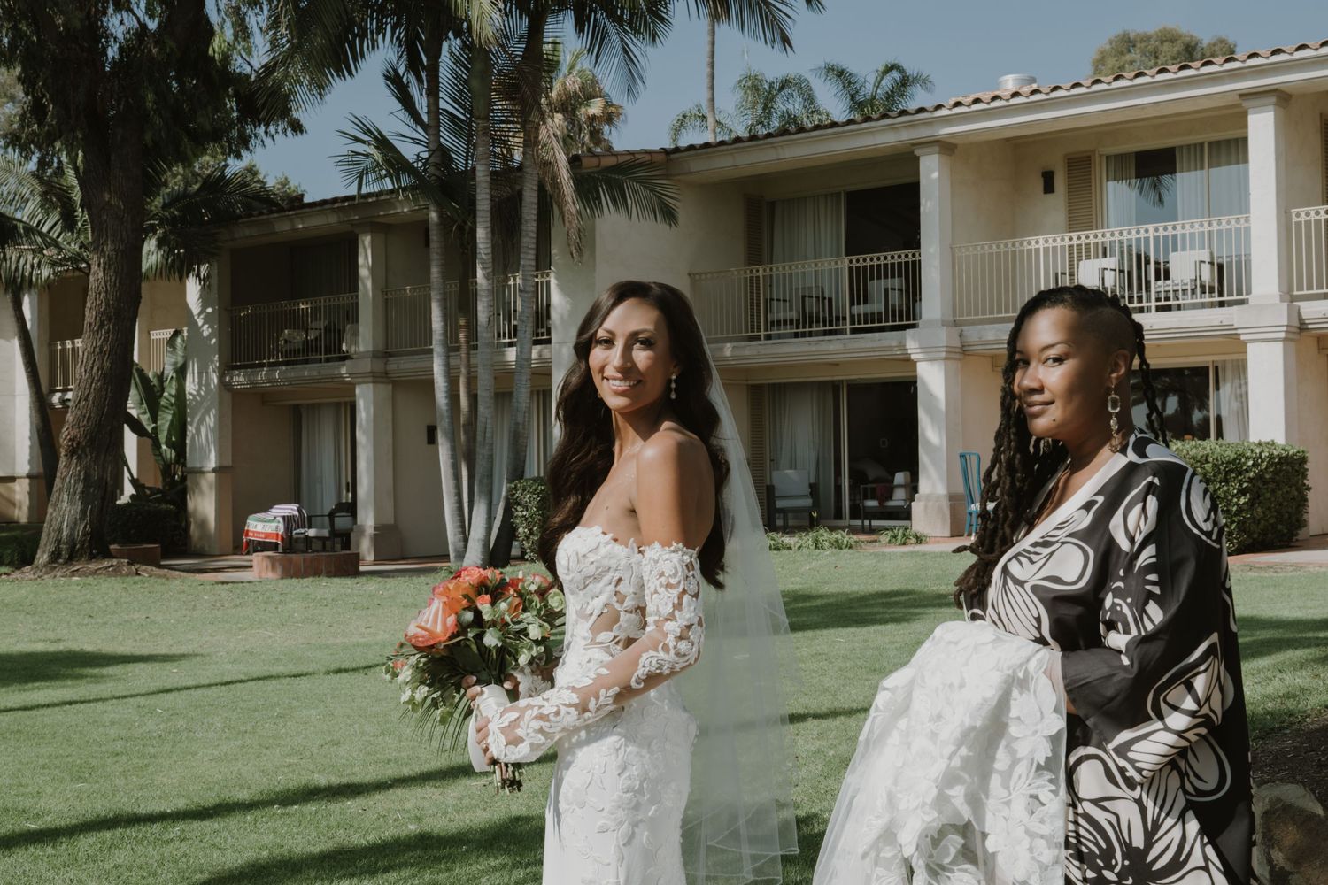 Wedding party poses on a lush green lawn with palm trees and Spanish-style buildings in the background.