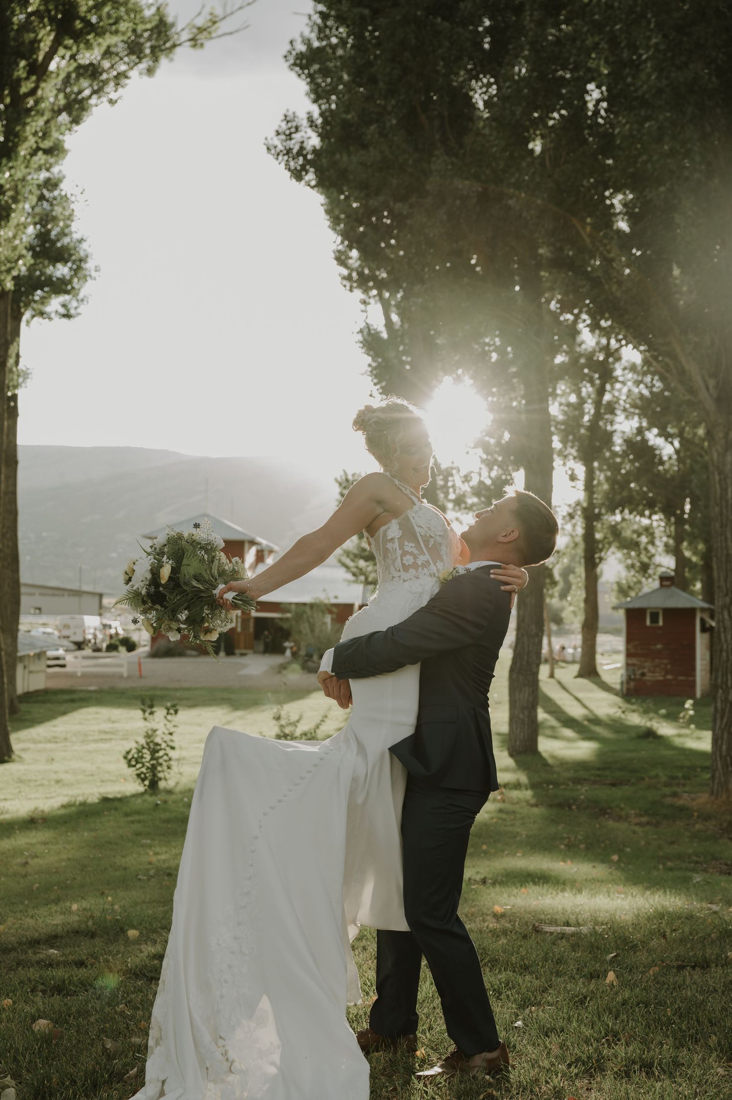 A wedding couple dances in golden sunlight filtering through trees near rustic buildings.