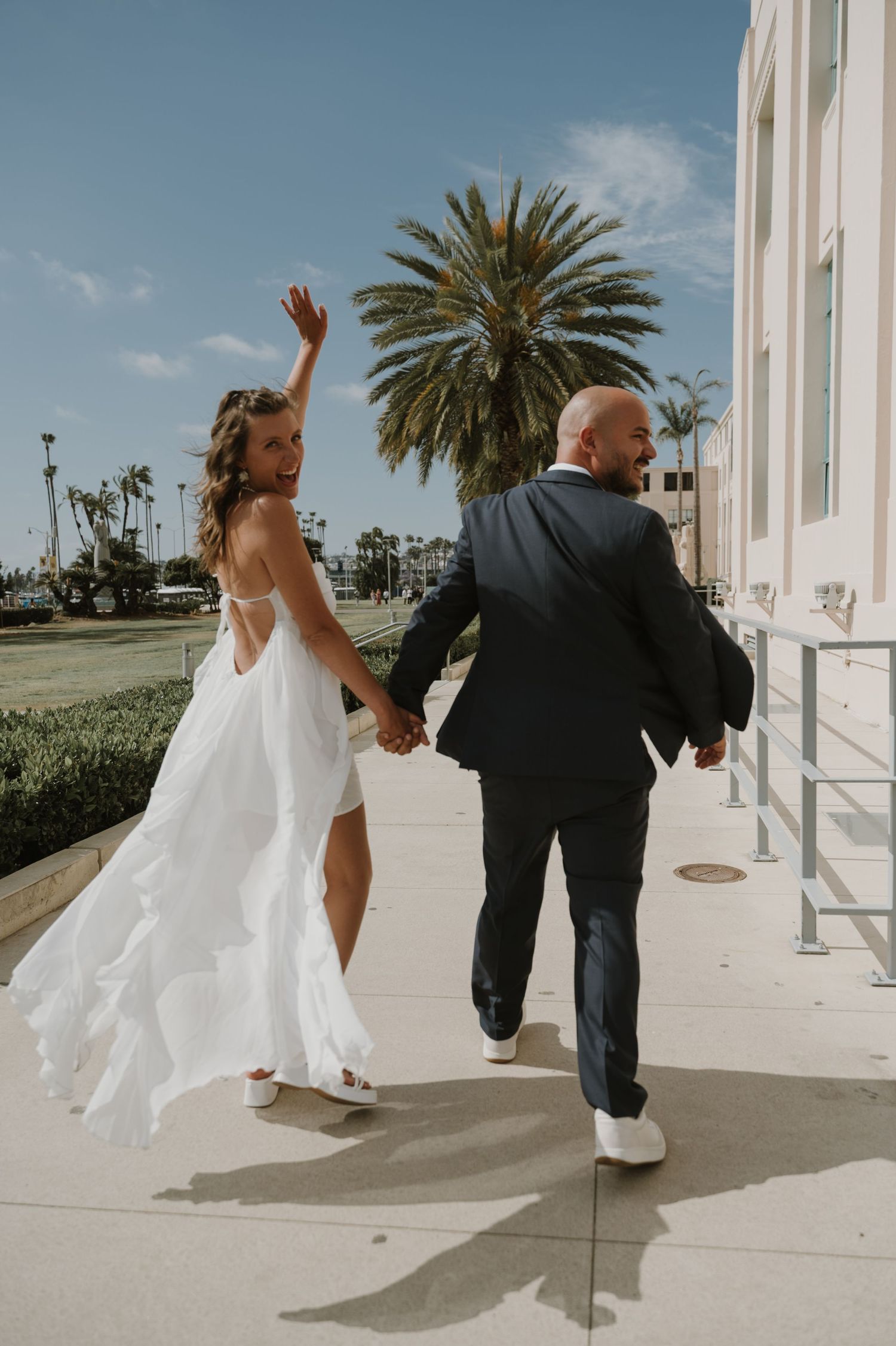 Couple in wedding attire walking and dancing outside a white building with palm trees.