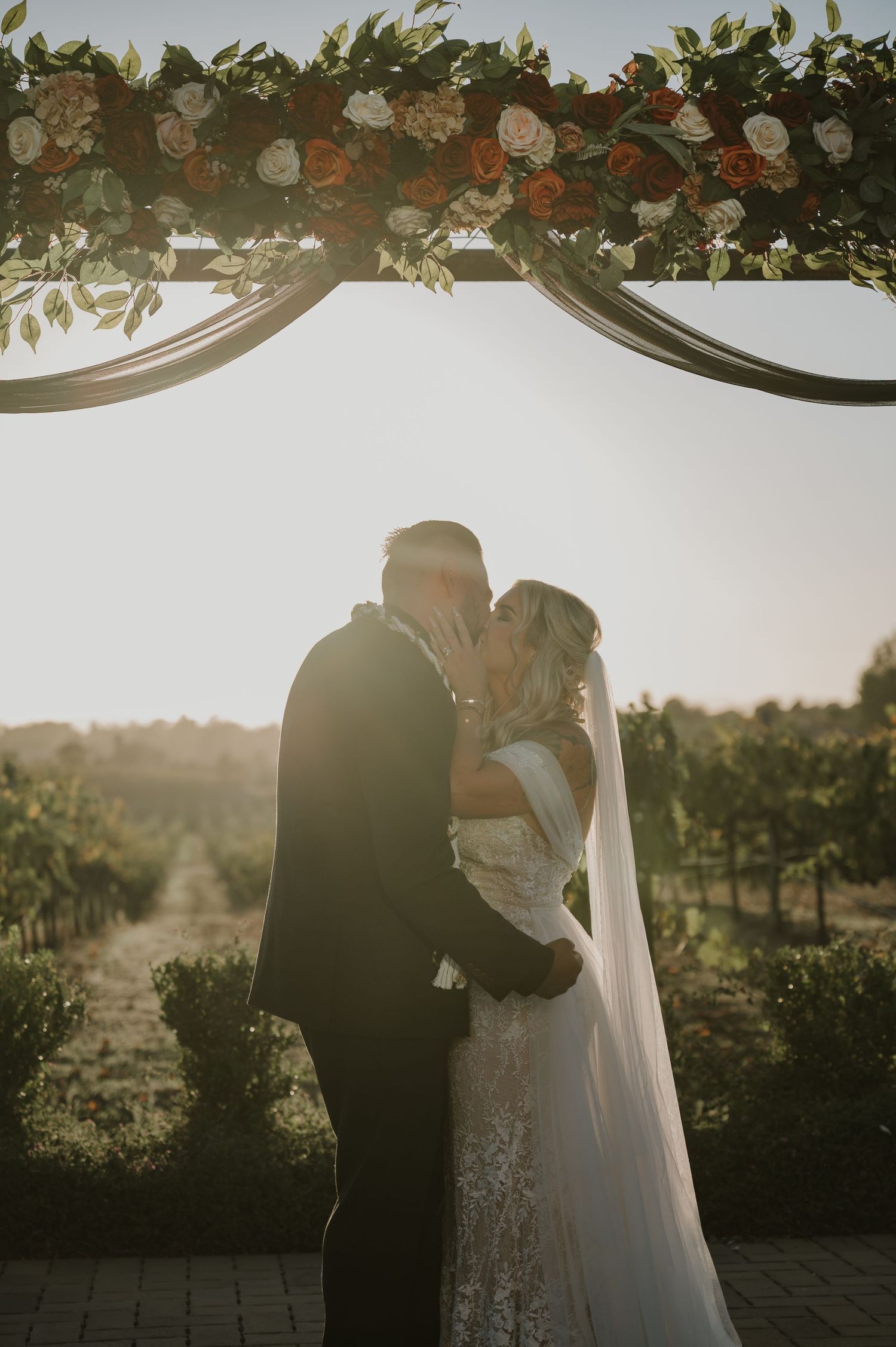 Silhouetted wedding couple sharing a kiss under floral arch with vineyard backdrop.