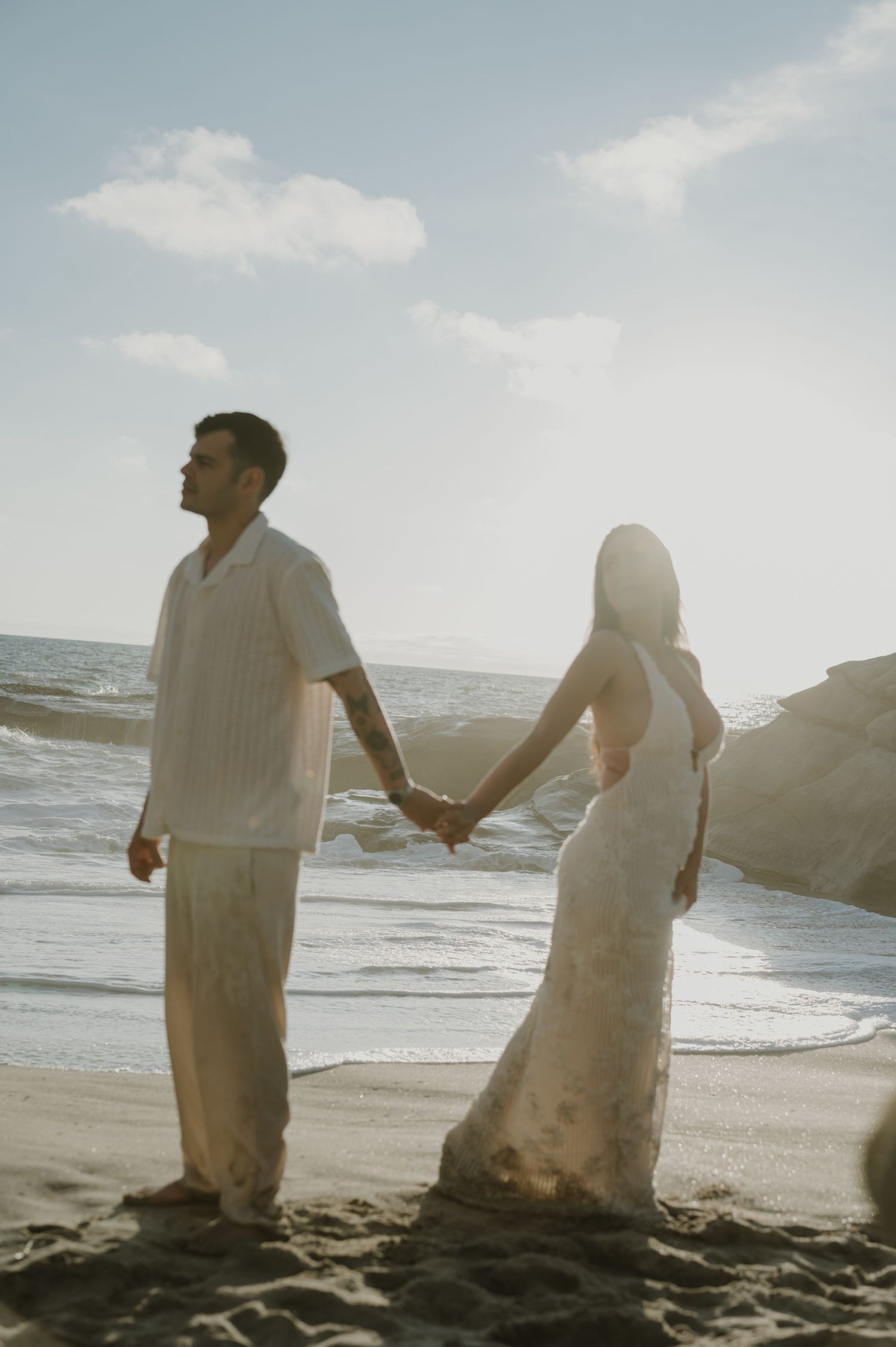 Couple walking along a sunlit beach shore holding hands in white flowing outfits.