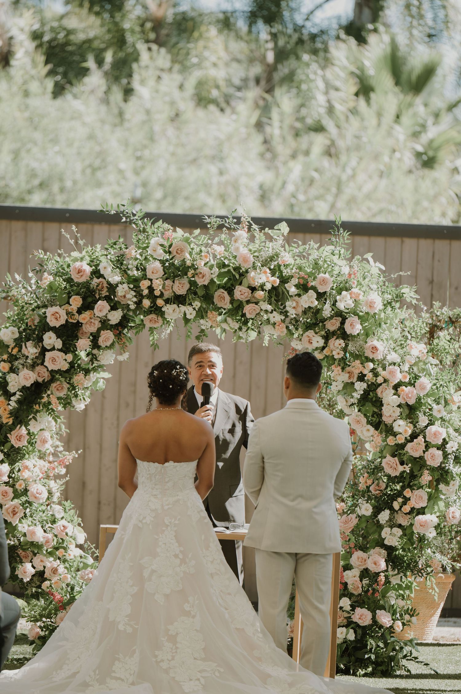 Bride and groom stand before a floral arch of blush roses during an outdoor wedding ceremony.