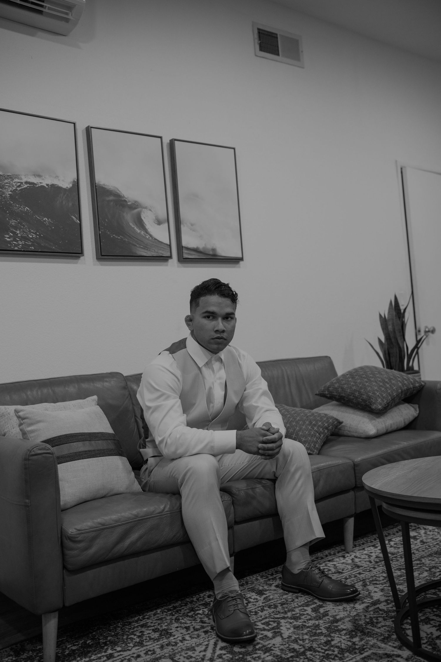Groom in white suit sitting on leather couch with ocean wave artwork behind him, black and white photo.