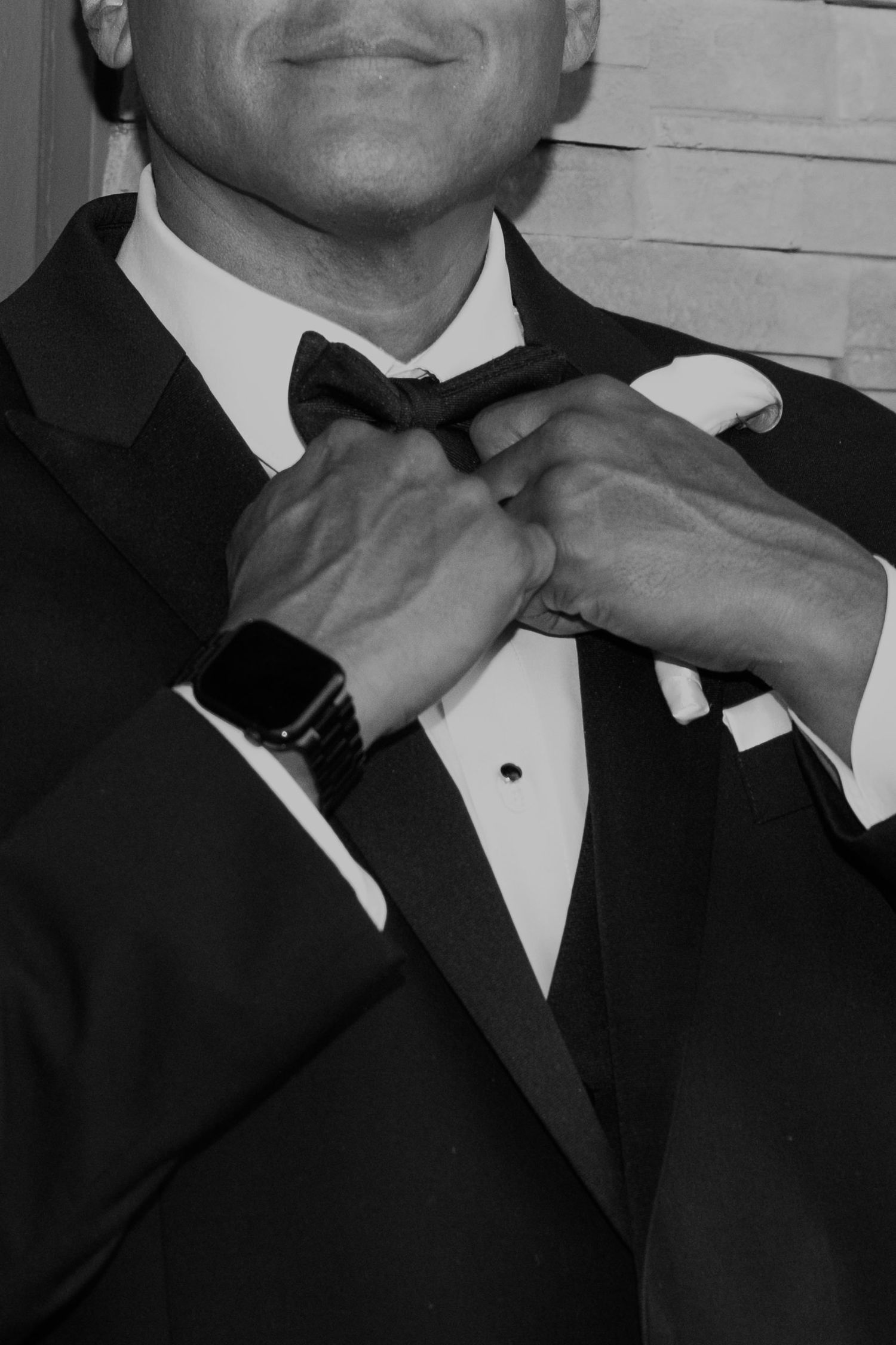 Man in tuxedo adjusting bow tie, wearing a smartwatch, black and white formal portrait photo.