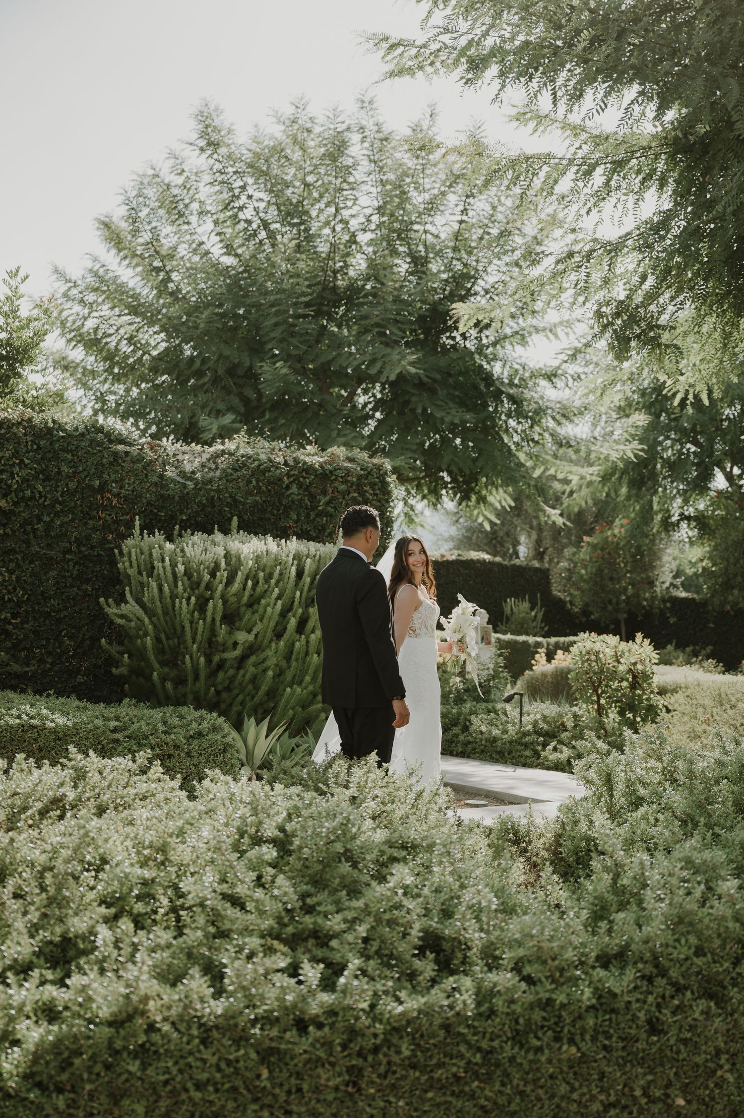 Bride and groom stand together in a lush garden surrounded by manicured hedges and tall trees on their wedding day.