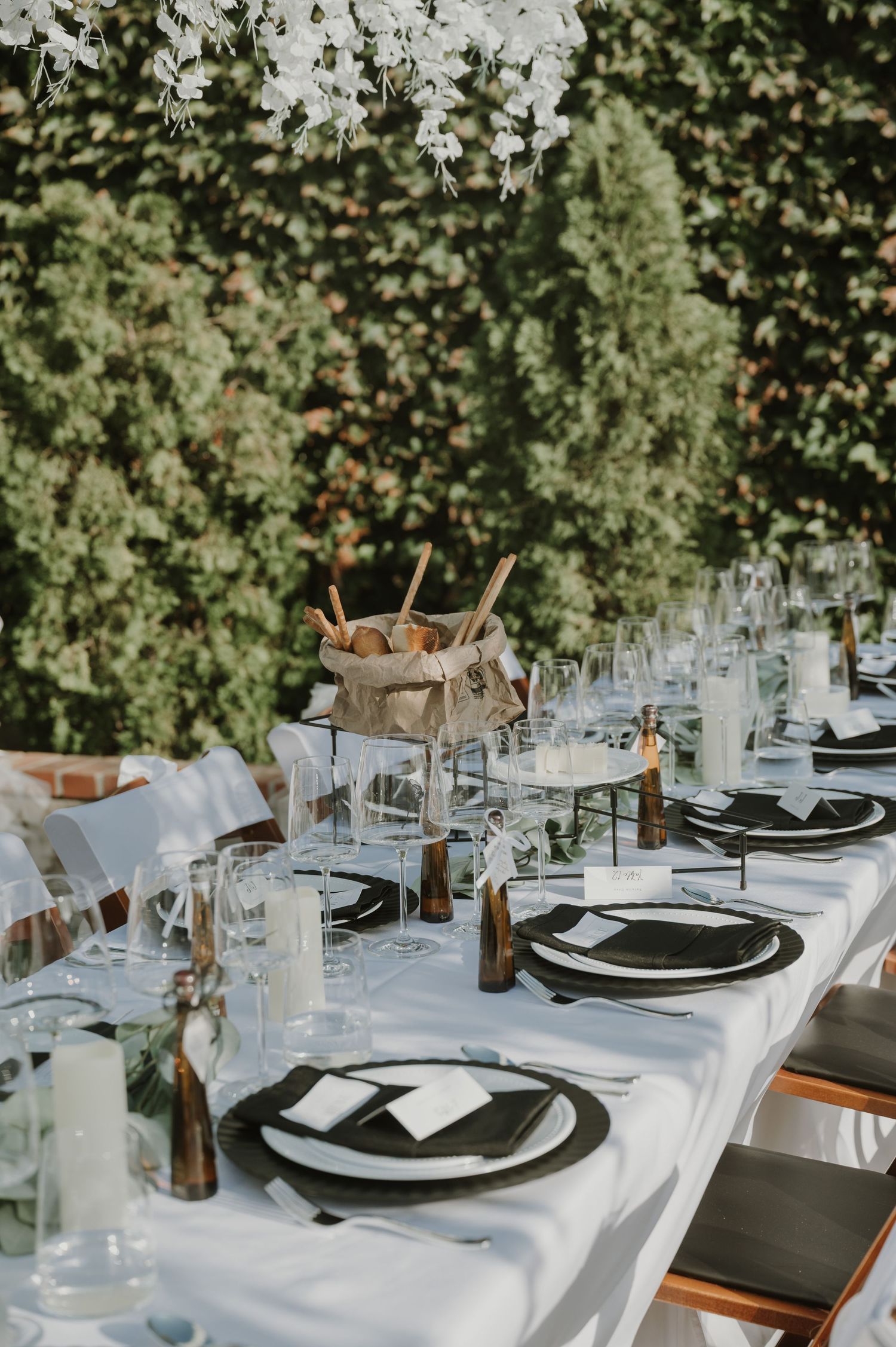 Sophisticated wedding reception table setting with black plates, crystal glasses, white linens, and lush green wall backdrop.