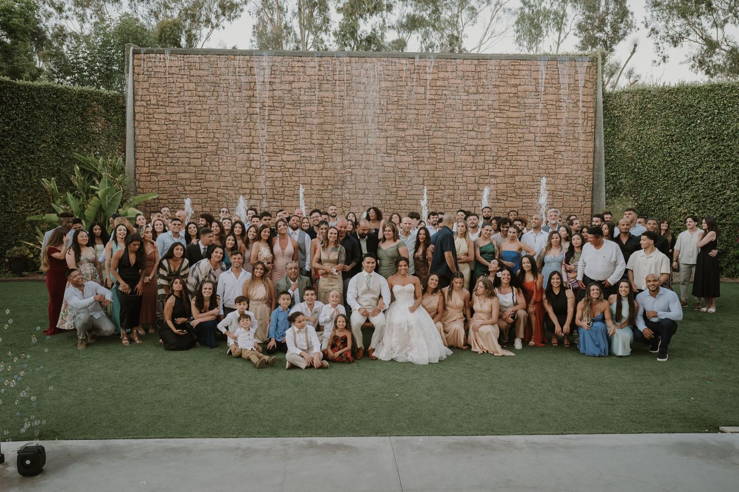 Large wedding group photo with all guests posing in front of decorative brick wall in garden venue.