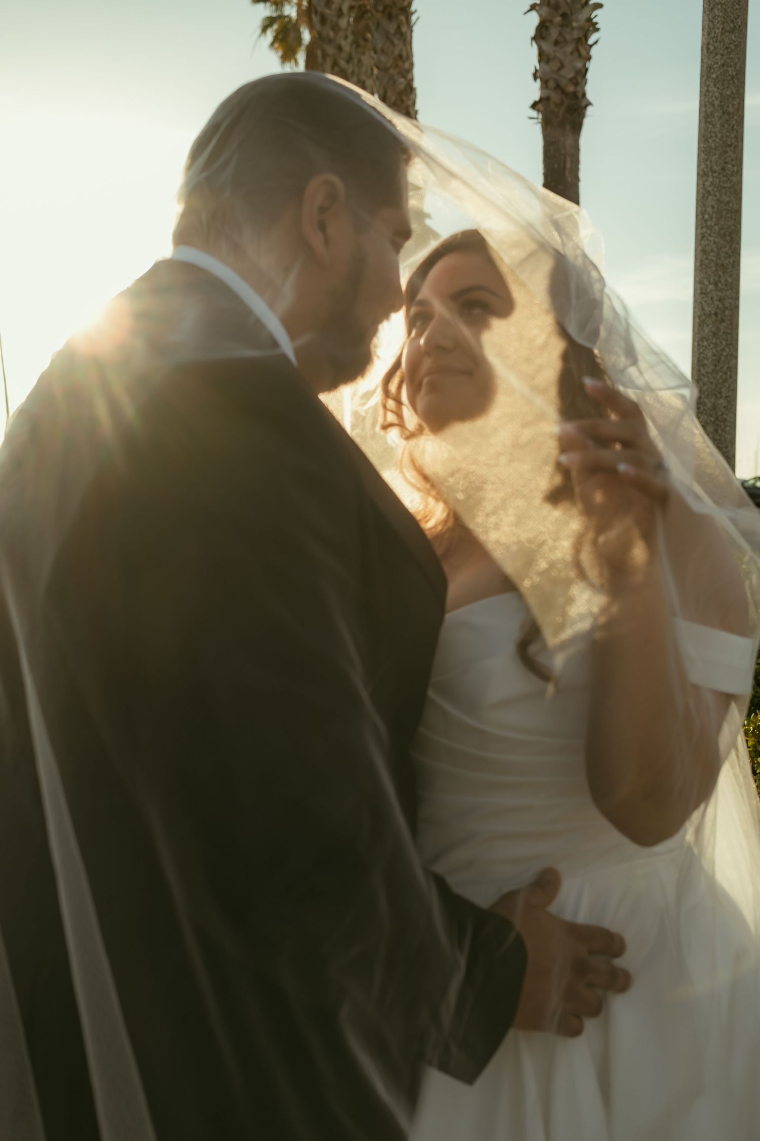 Bride and groom share a romantic moment under a veil at golden hour, with palm trees in the background.