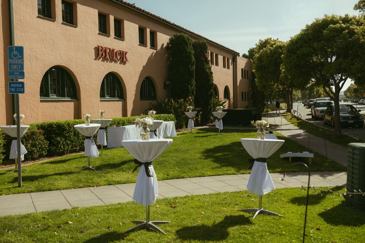 Outdoor wedding cocktail hour setup with white draped bistro tables on green lawn in front of a tan building.