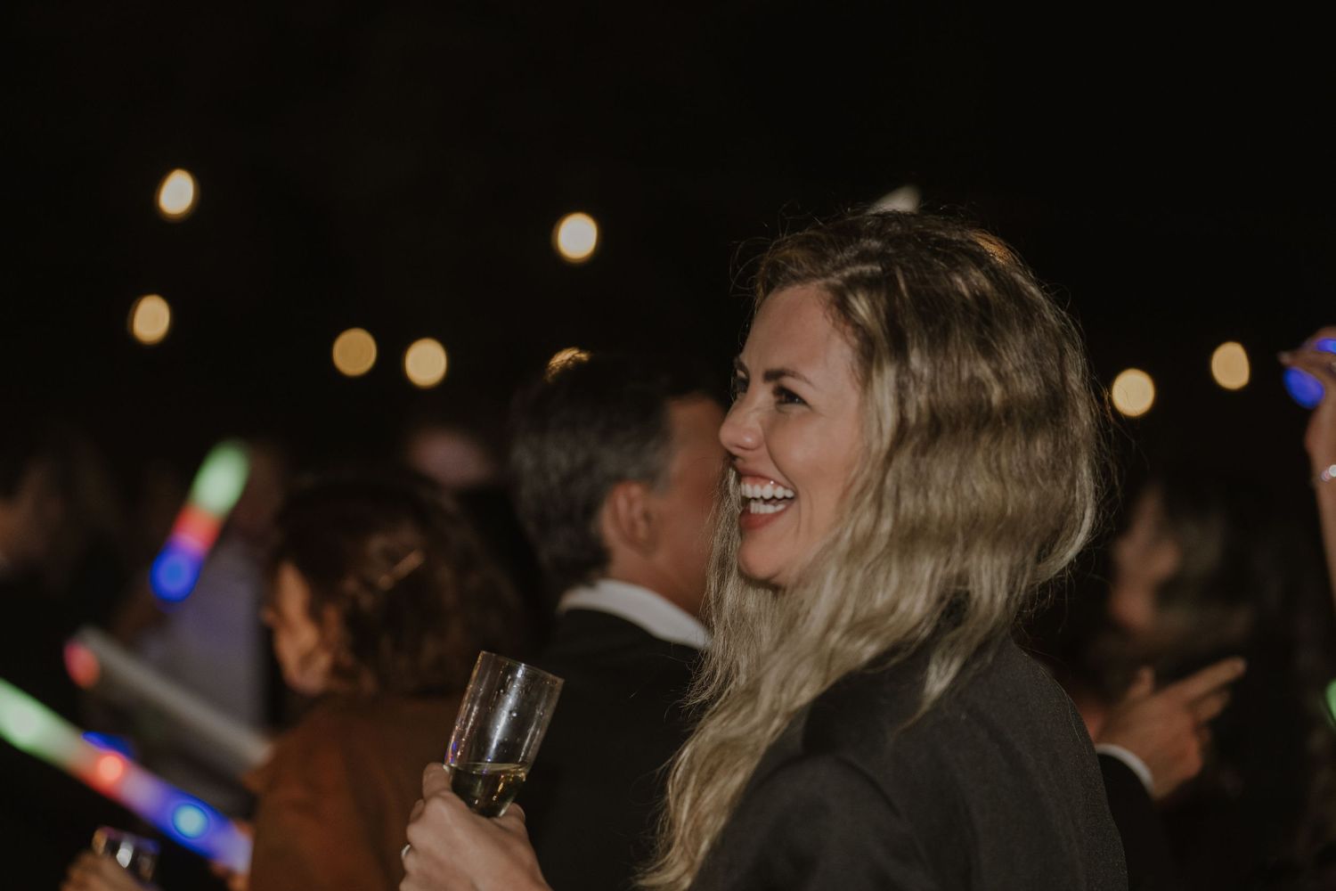 Smiling blonde woman holding a champagne glass at an outdoor evening wedding reception.