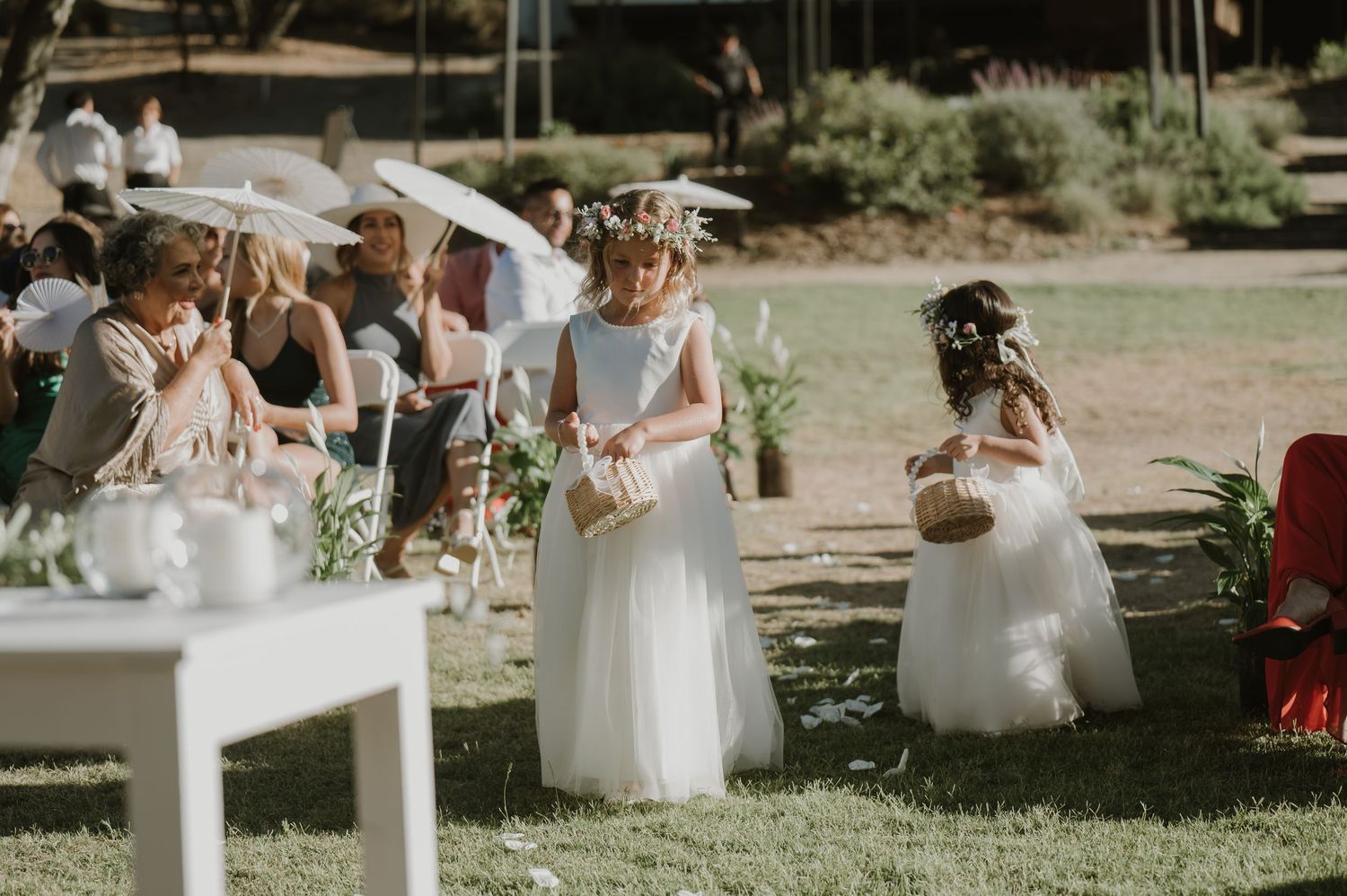 Two flower girls in white dresses walk down outdoor wedding aisle scattering petals.