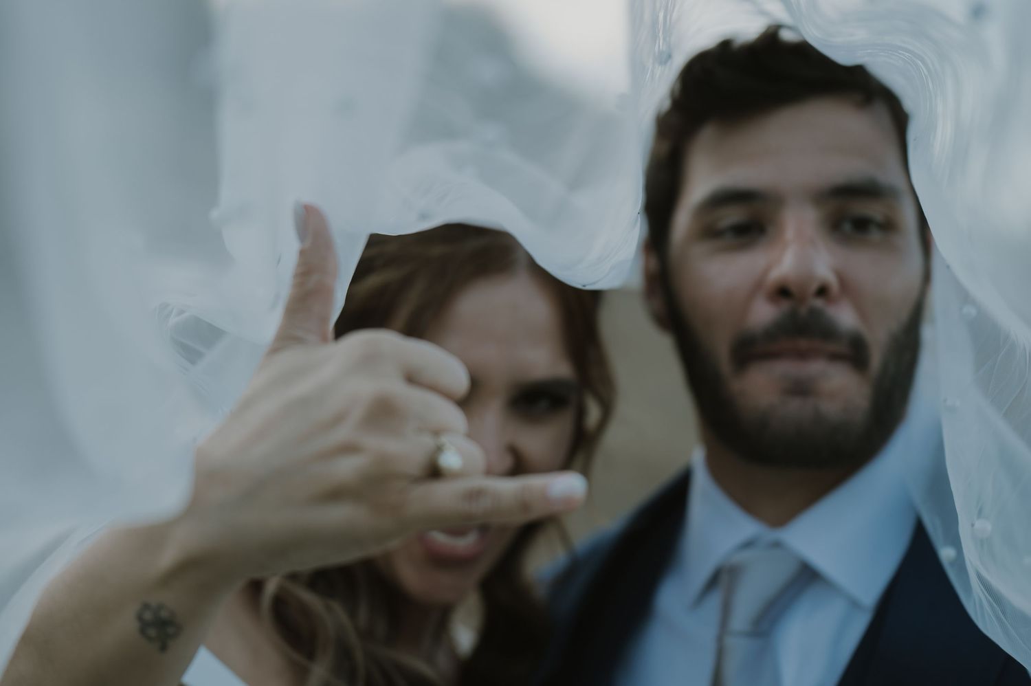 Bride and groom pose together showing off wedding ring, with soft white veil in background.