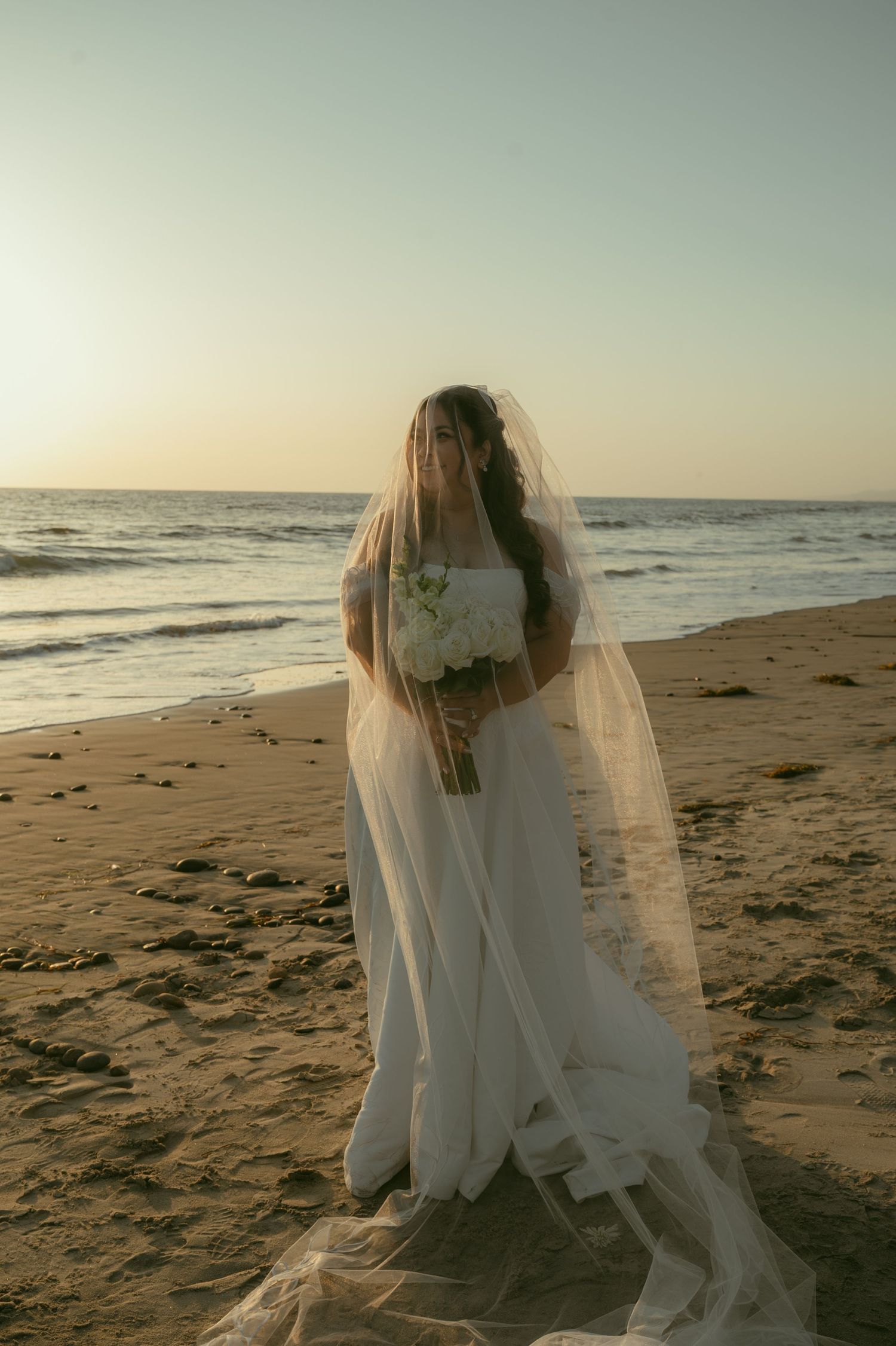 Bride in white gown and veil holding bouquet on sandy beach at sunset with ocean waves in background.