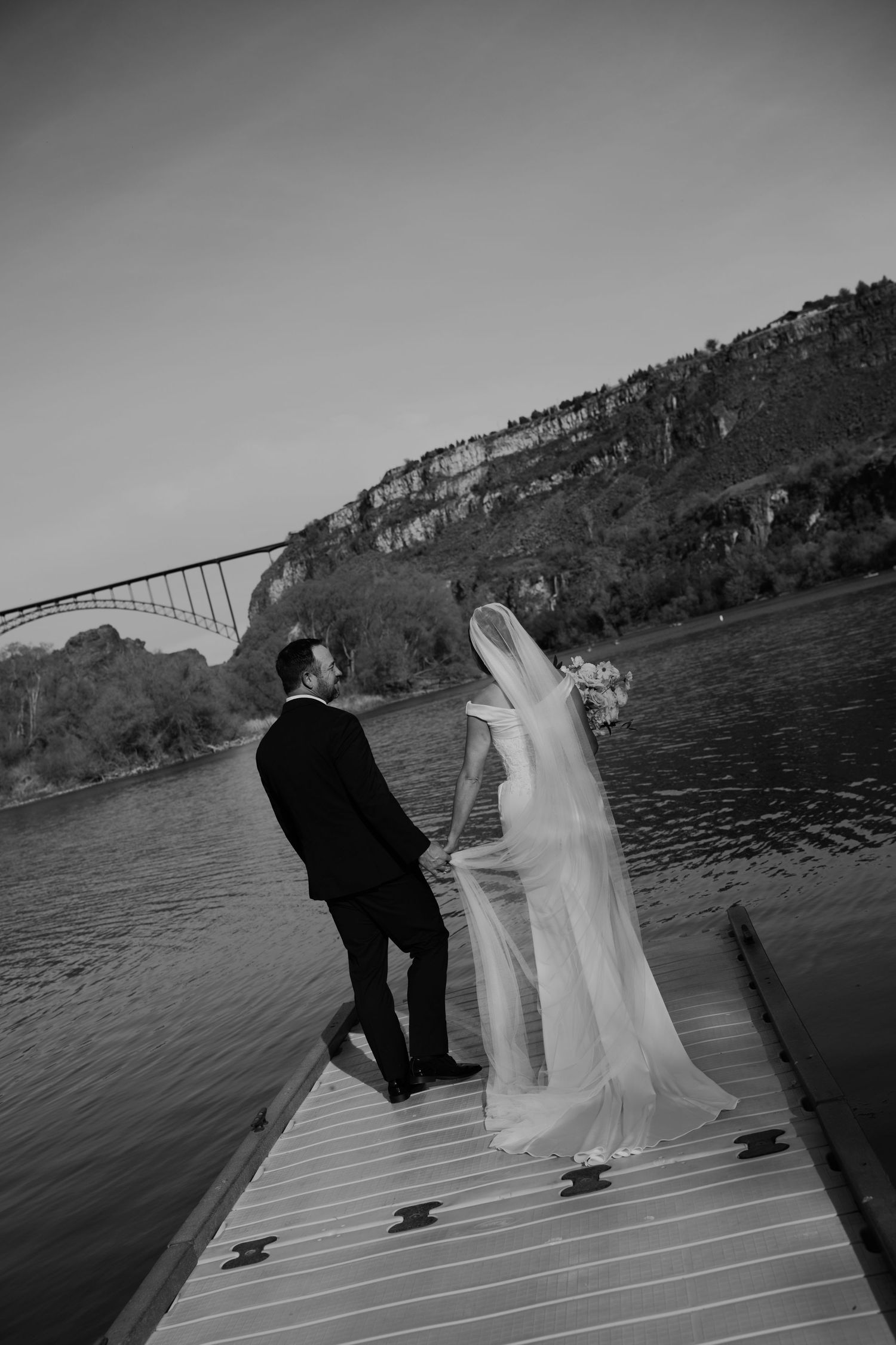 Black and white photo of bride and groom holding hands on a dock by a river with a bridge and cliffs in the background.