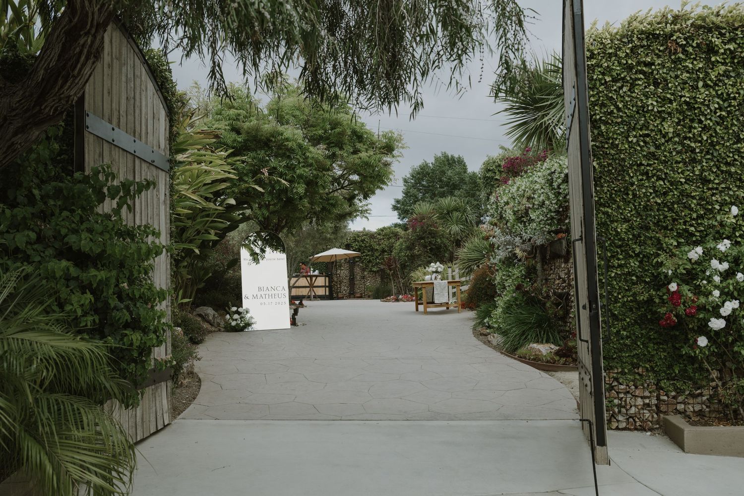 Lush garden venue entrance with tall hedges, tropical plants, and a winding driveway leading to an outdoor event space.