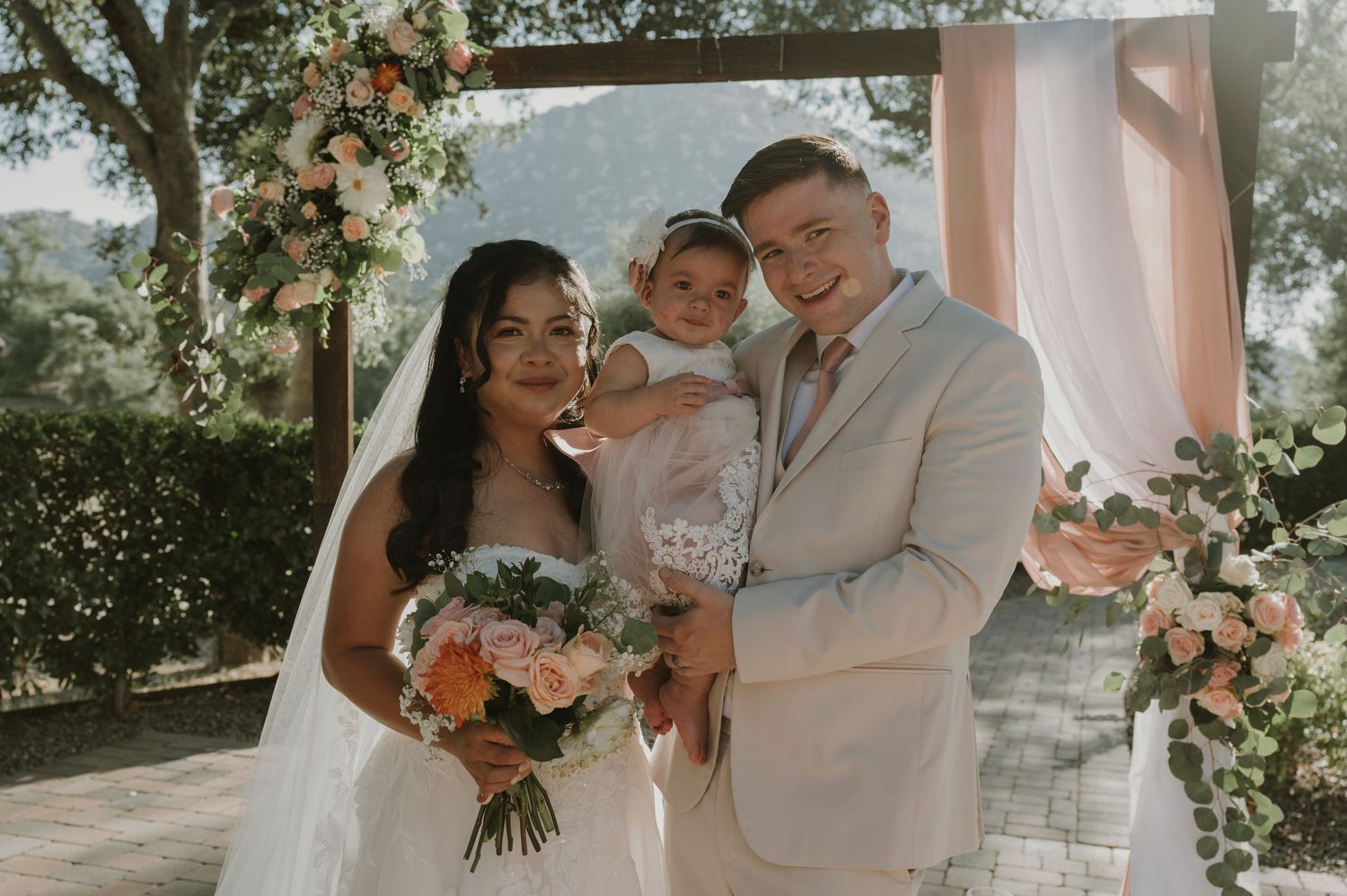 Newlyweds pose with their young child under a floral arch at their outdoor wedding ceremony.
