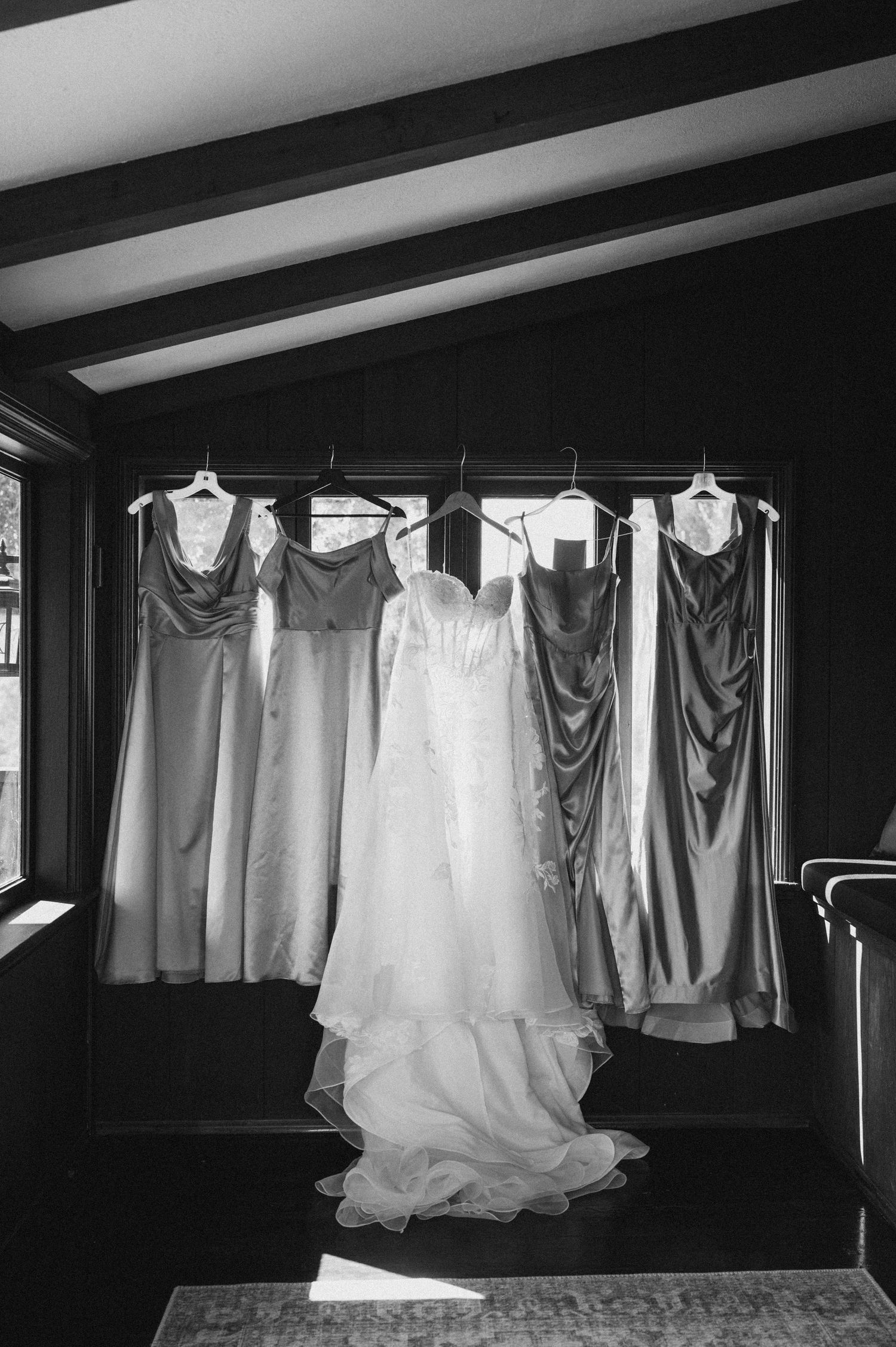 Black and white photo of a wedding gown and four bridesmaid dresses hanging in a row near a window.