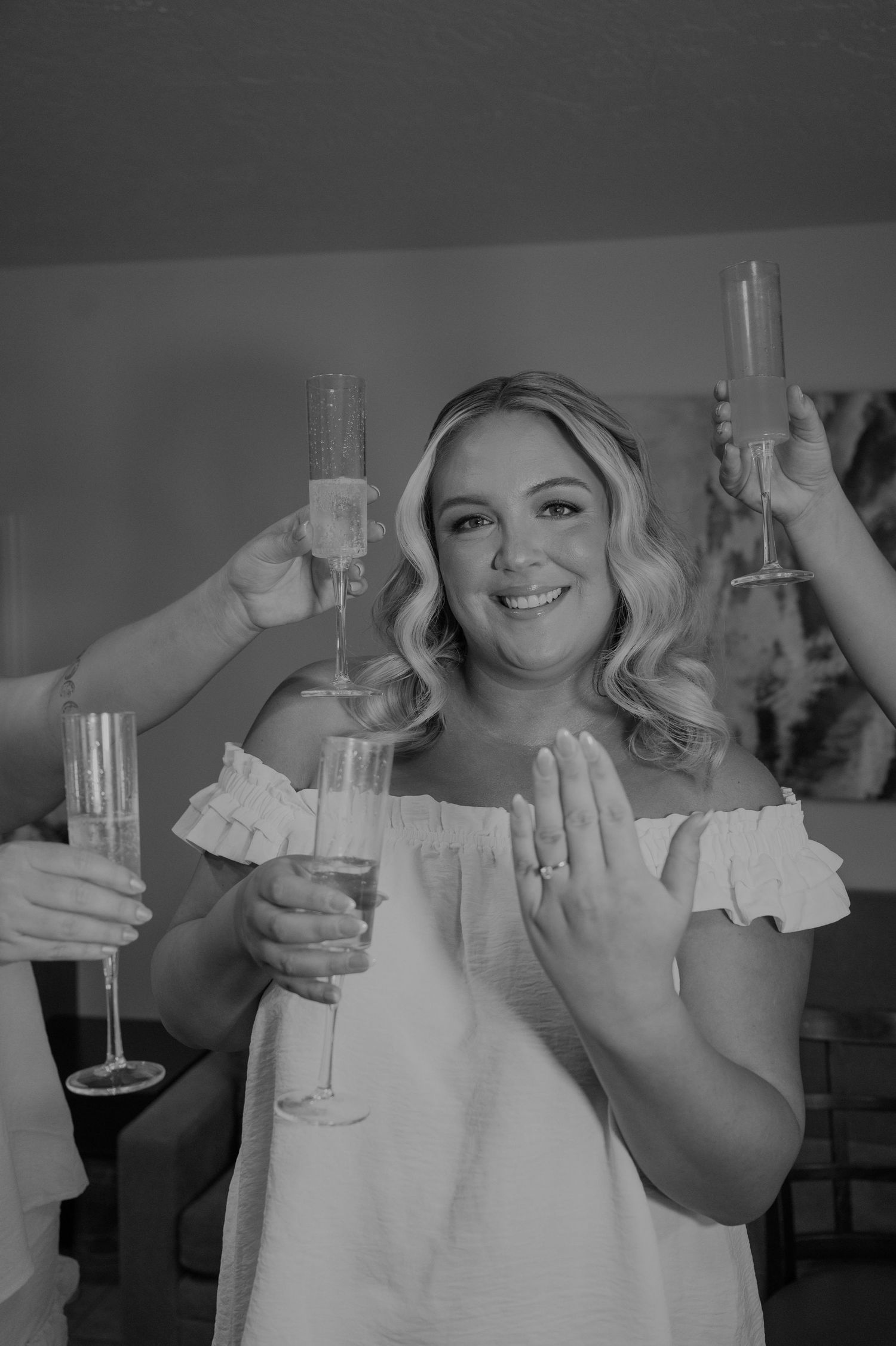 Bride smiling and showing engagement ring while toasting champagne flutes with bridesmaids during wedding preparations.