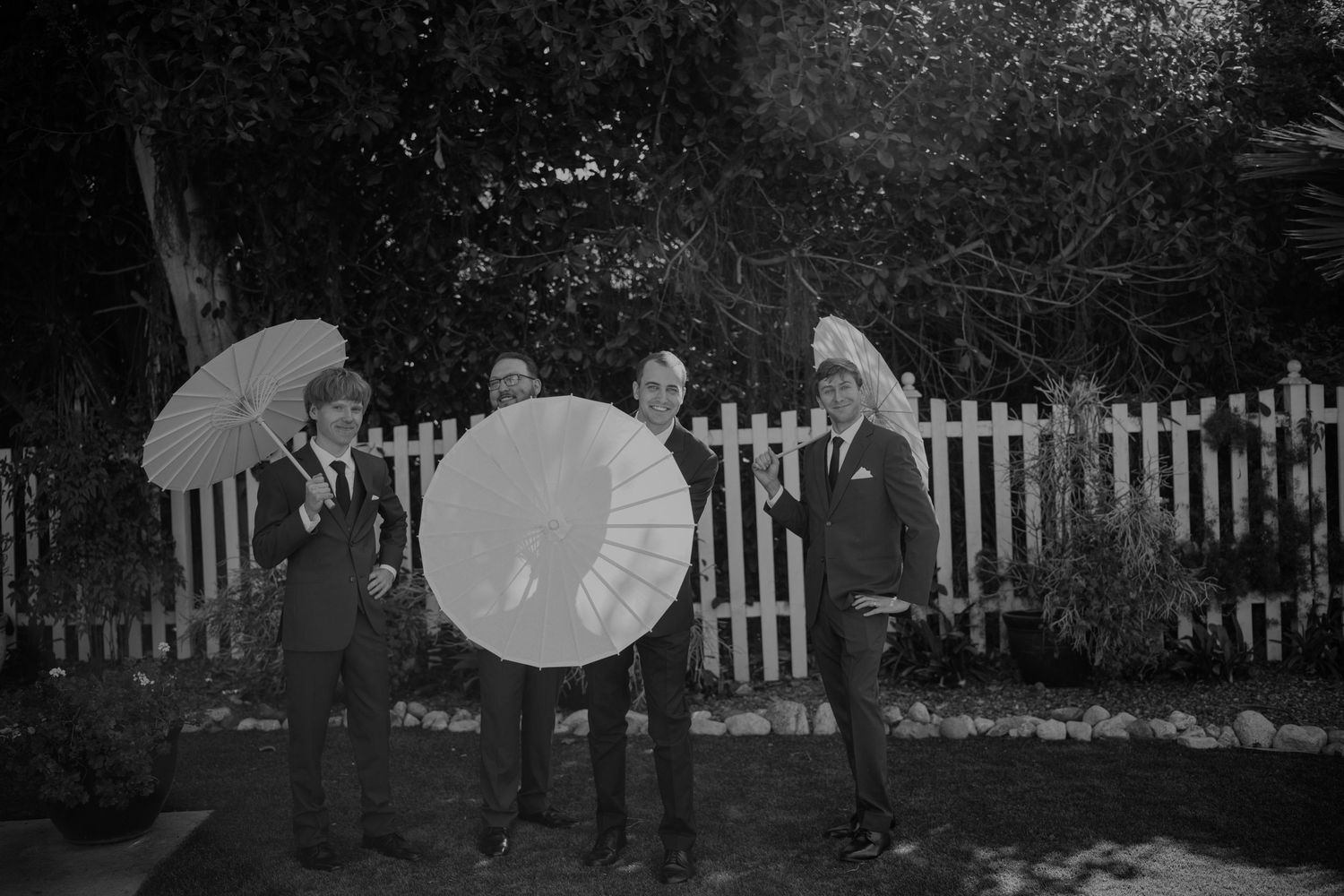Three men in suits holding decorative parasols outdoors near a white picket fence in a black and white photo.