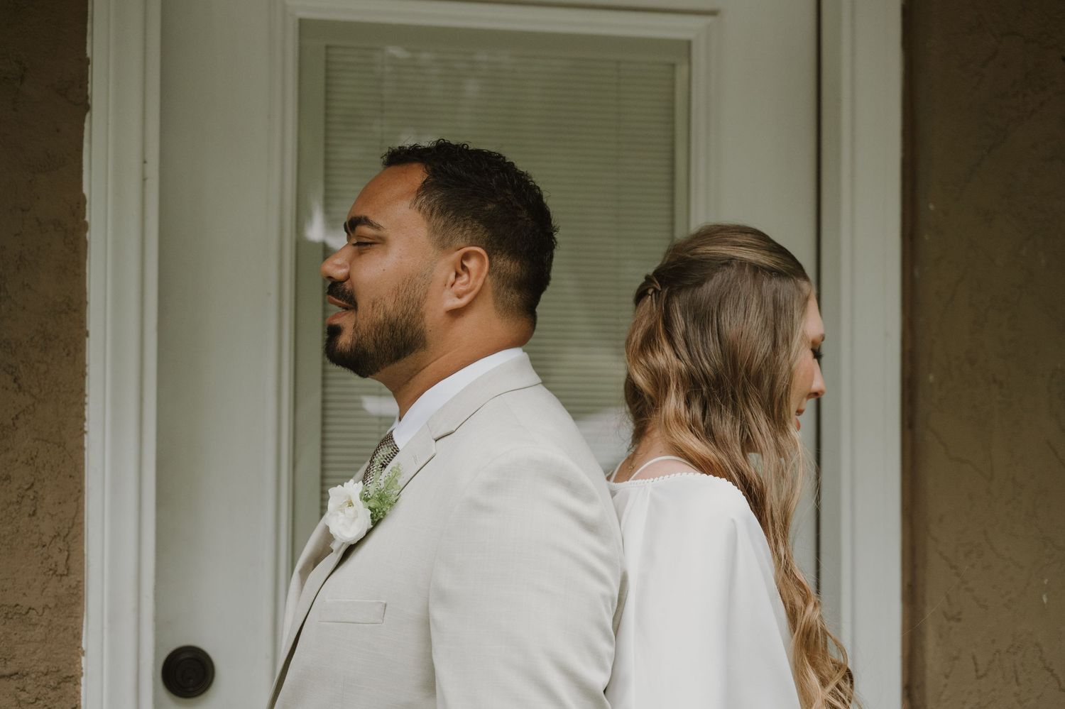 A bride and groom stand back-to-back in a first look moment outside a white door before their wedding ceremony.