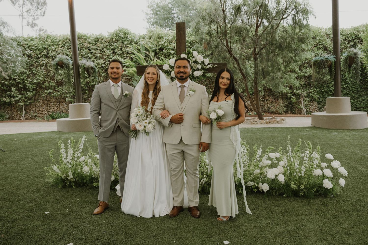 Wedding party of four posing outdoors on green lawn with white floral arrangements and wooden cross backdrop.