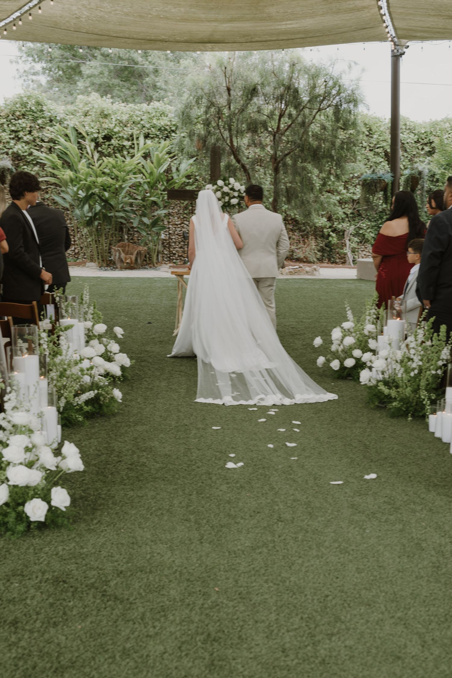 Bride walking down the aisle with white floral arrangements on green grass at an outdoor wedding ceremony.