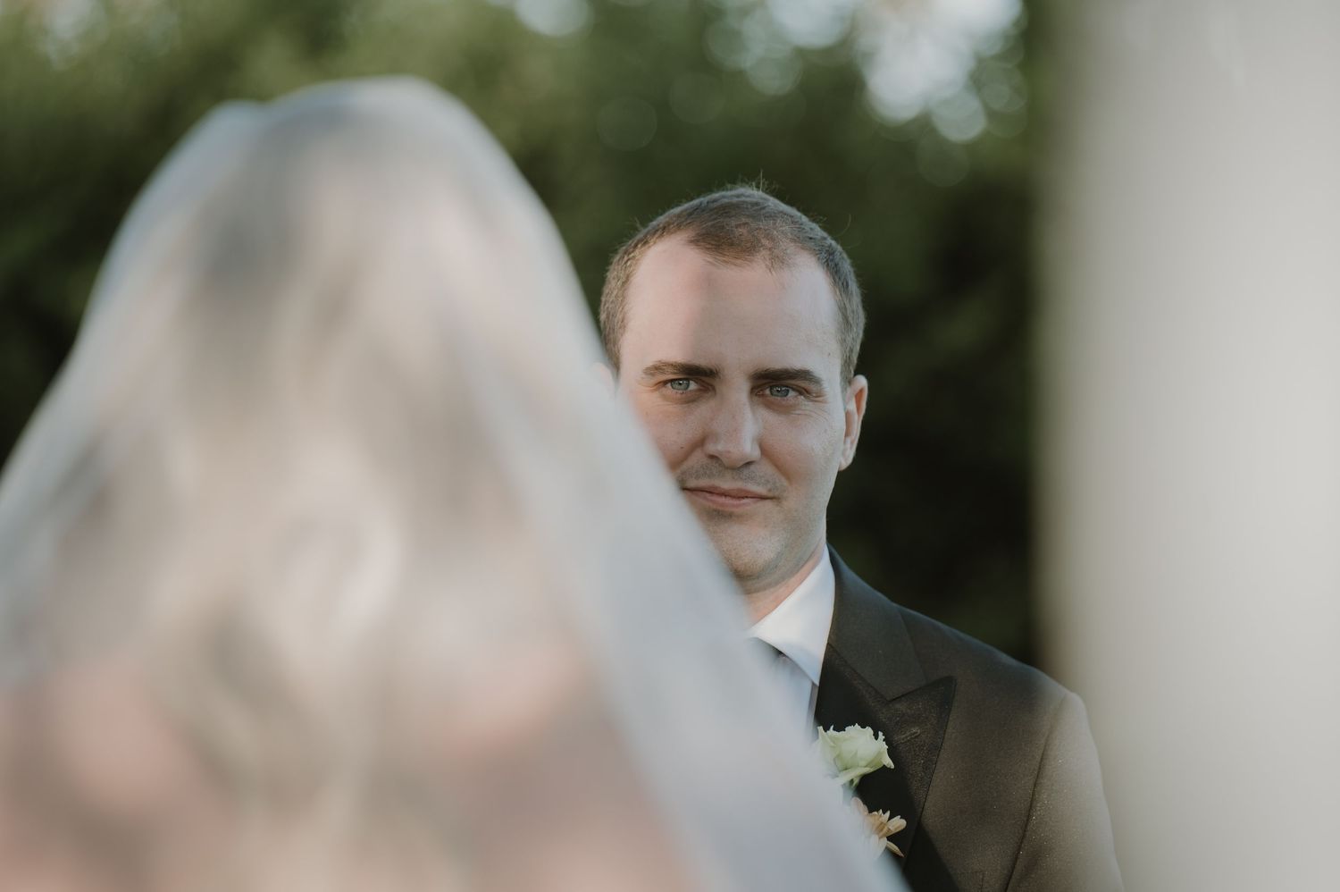 Groom in dark suit gazes lovingly at bride in wedding veil during outdoor ceremony.