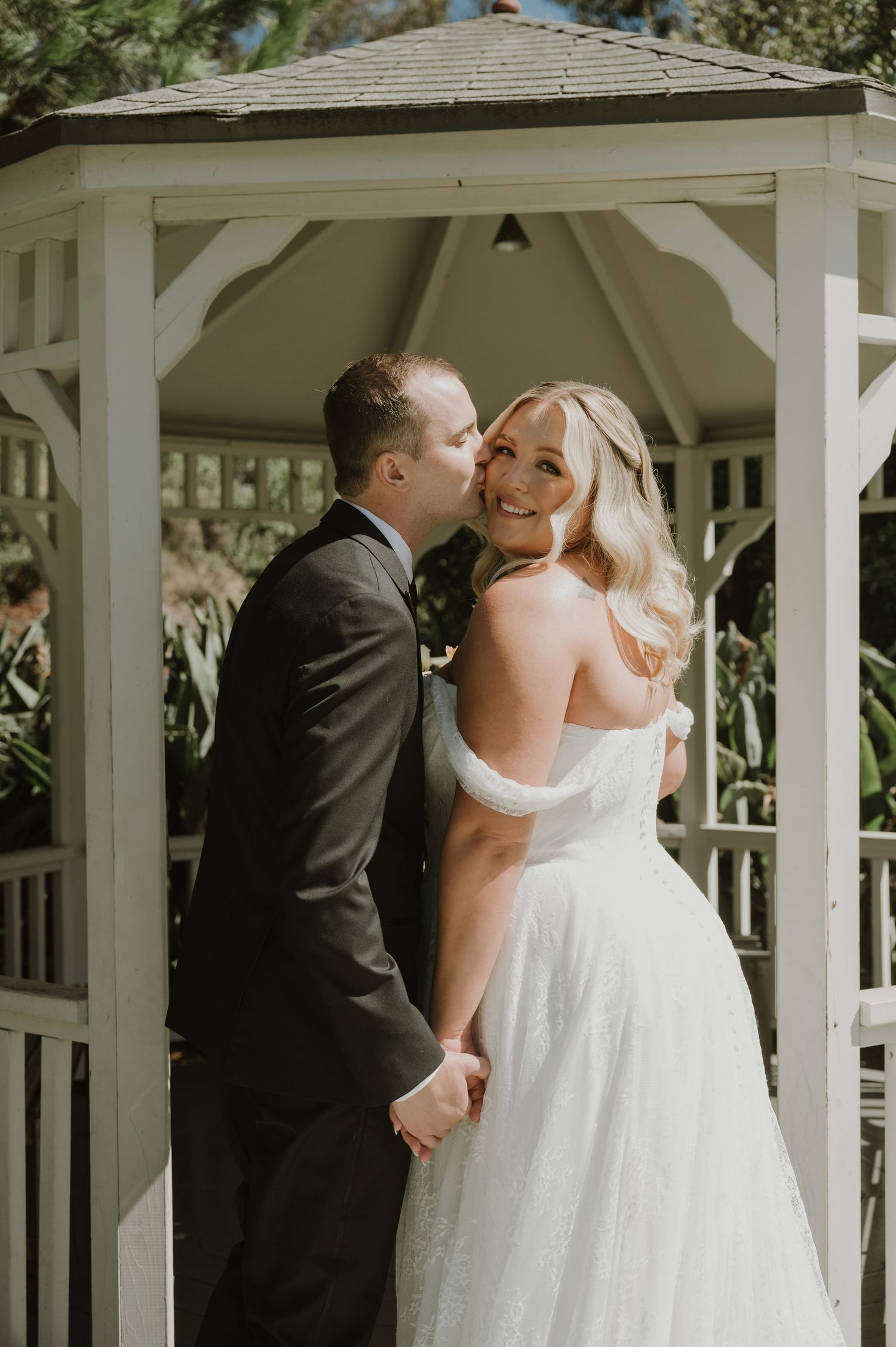 Groom in dark suit kissing bride in white off-shoulder gown under a white gazebo surrounded by lush greenery.