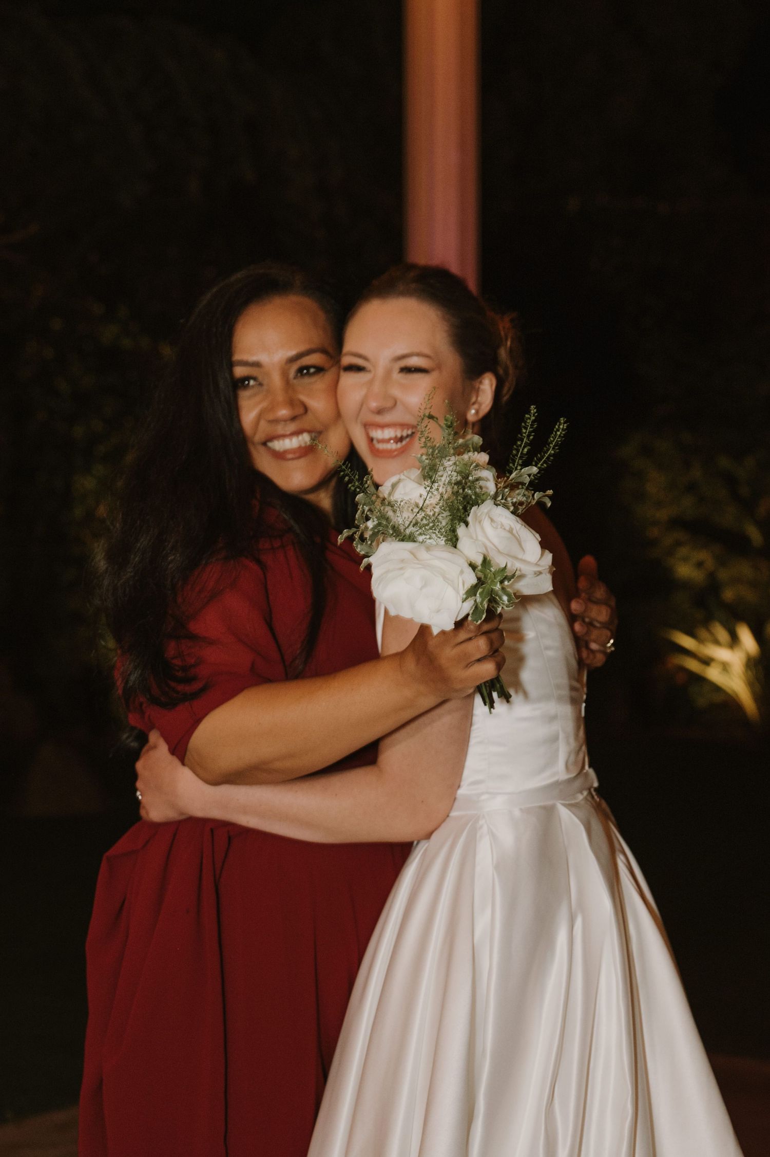 Bride in white satin gown laughing while embracing bridesmaid in red dress at night wedding.