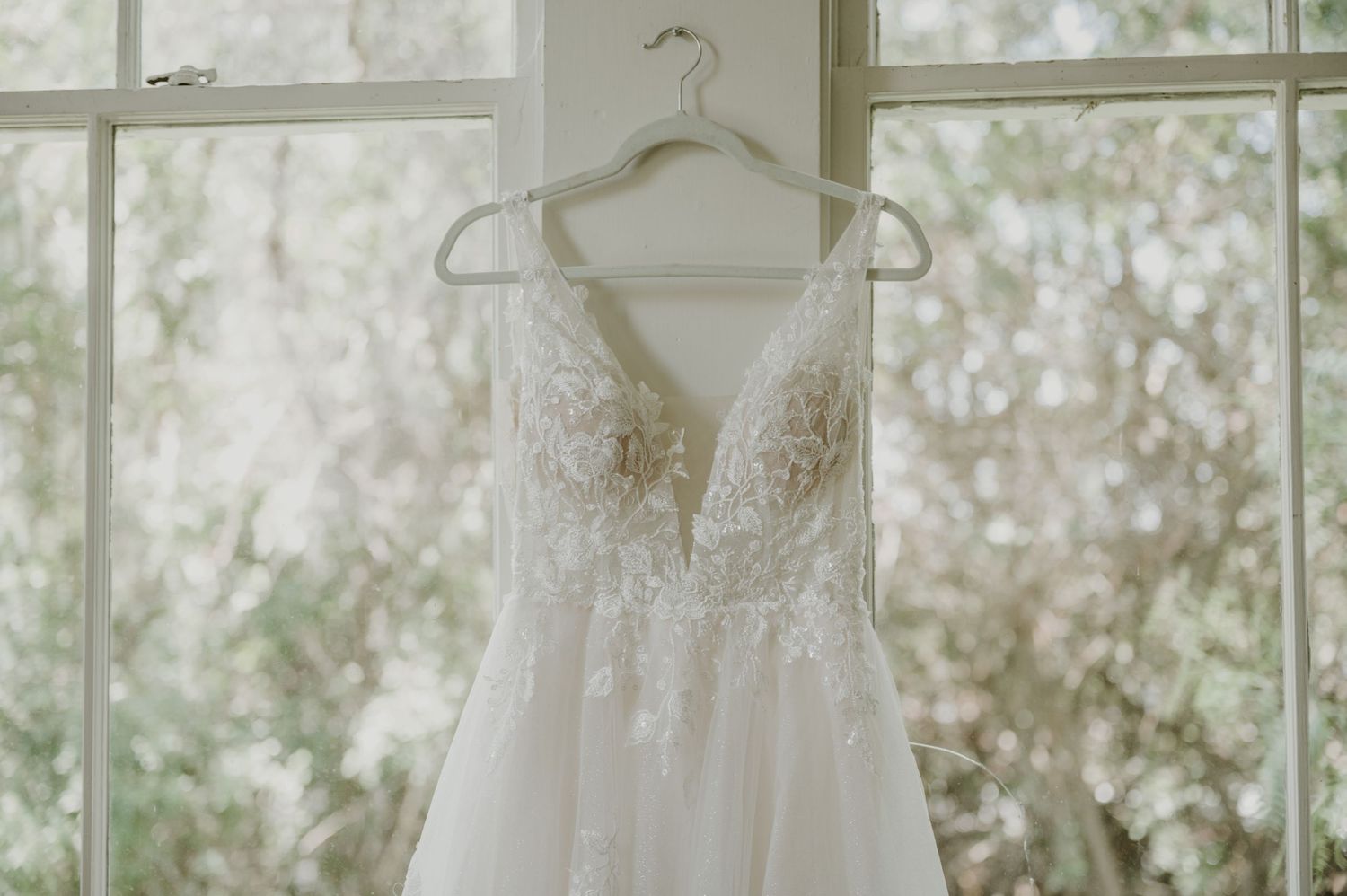 Elegant lace wedding dress with deep V-neckline hanging on a hanger in front of a bright window.