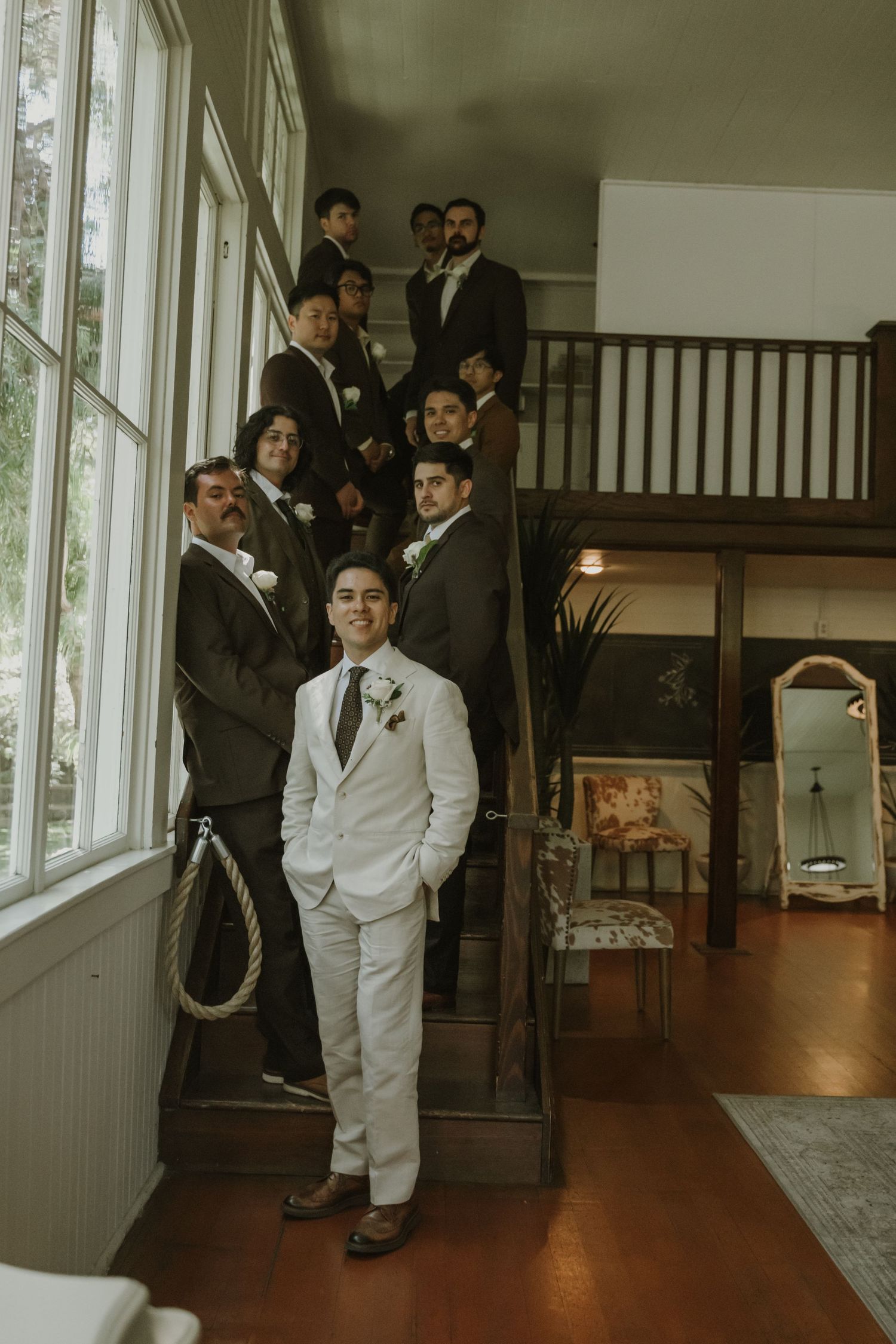 Groom in white suit poses with groomsmen in dark suits on elegant staircase.