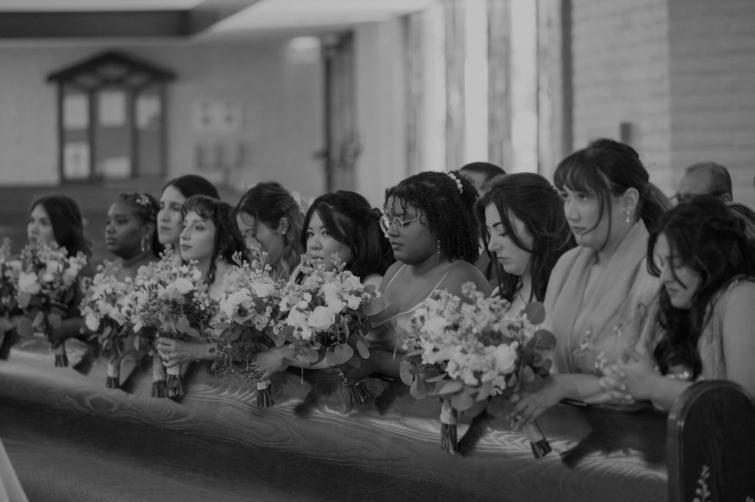 Bridesmaids holding floral bouquets seated in church pews in black and white.