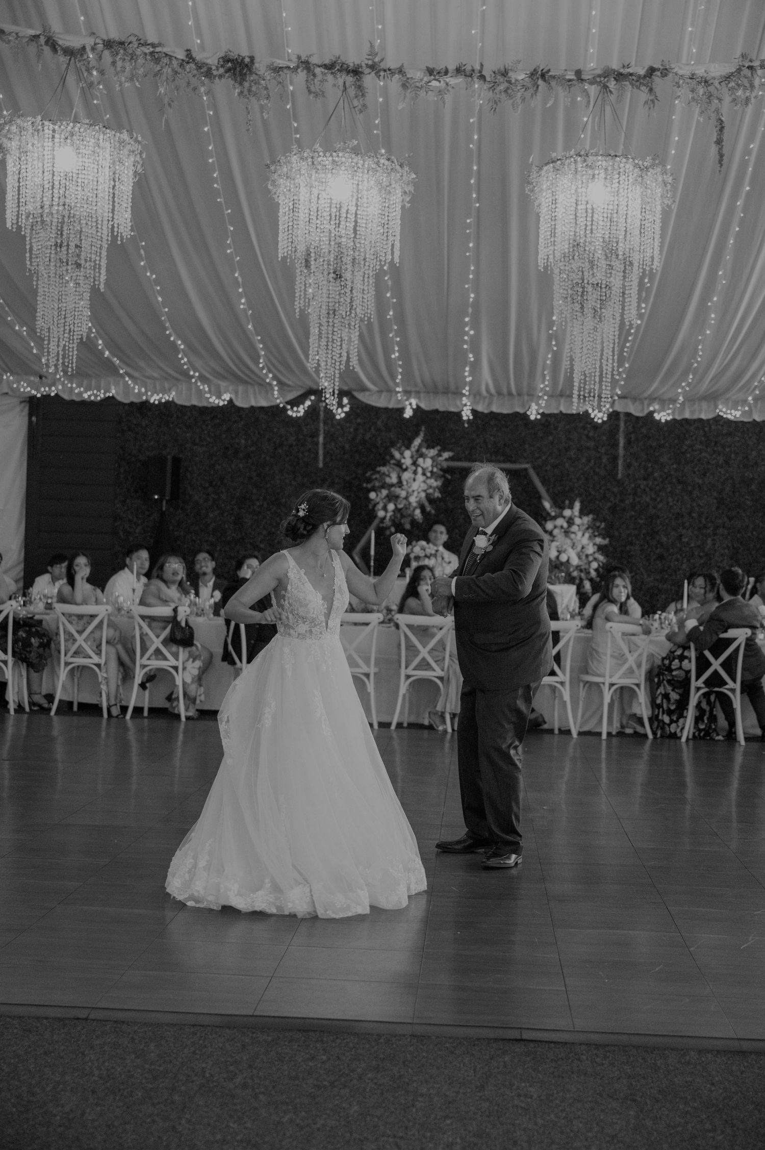 Bride and father share first dance at elegant wedding reception with crystal chandeliers and draped ceiling.