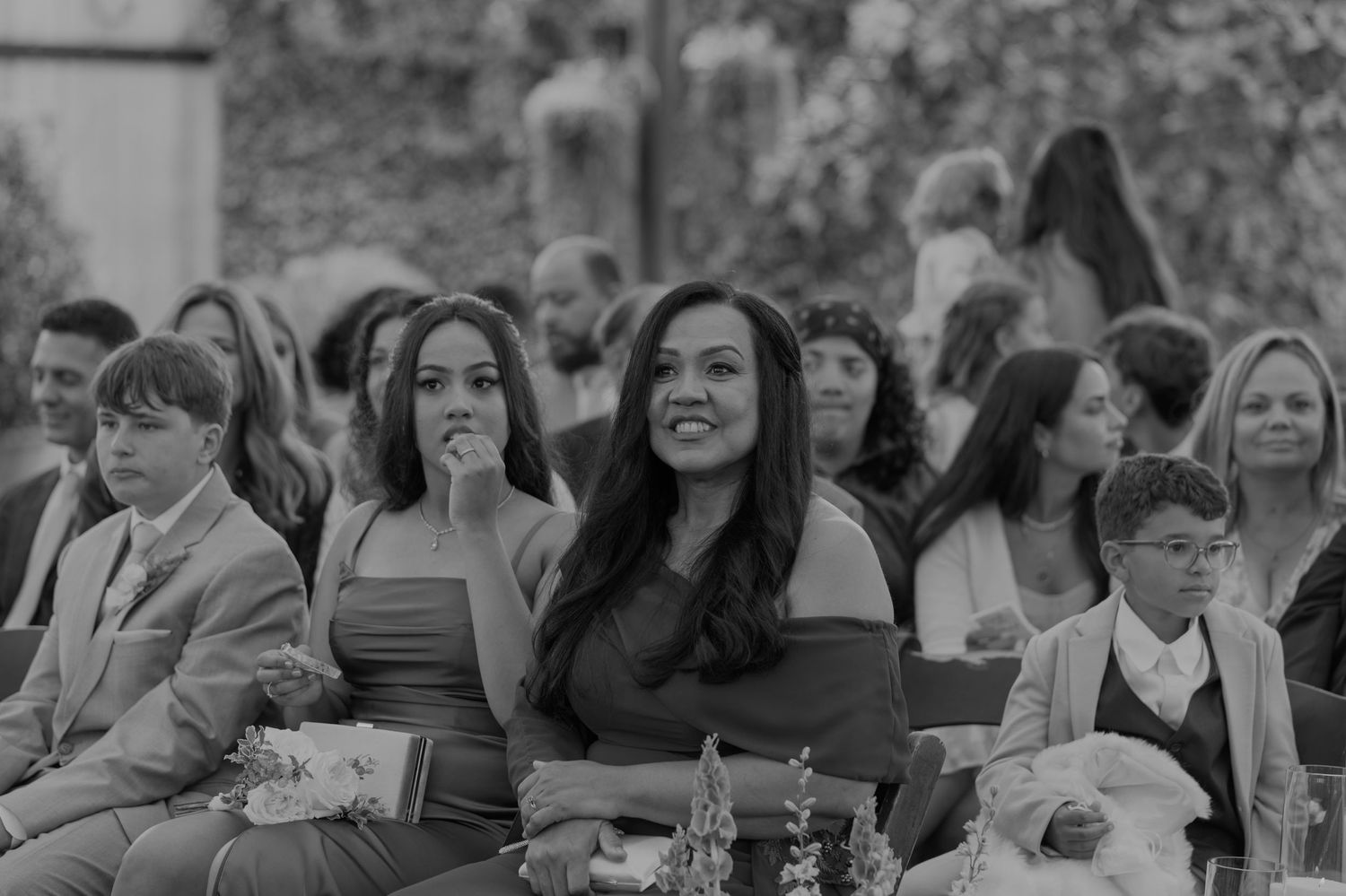Black and white photo of smiling wedding guests seated outdoors, enjoying a ceremony celebration.