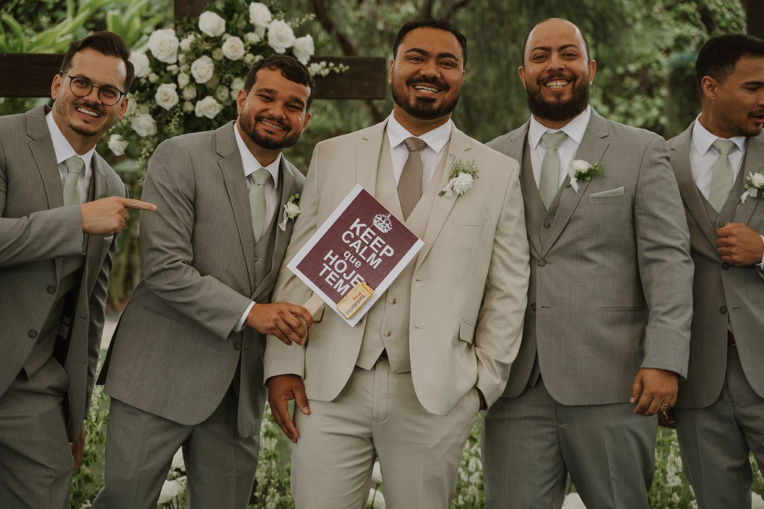 Groom and groomsmen in gray suits smile holding a humorous sign at an outdoor wedding ceremony.