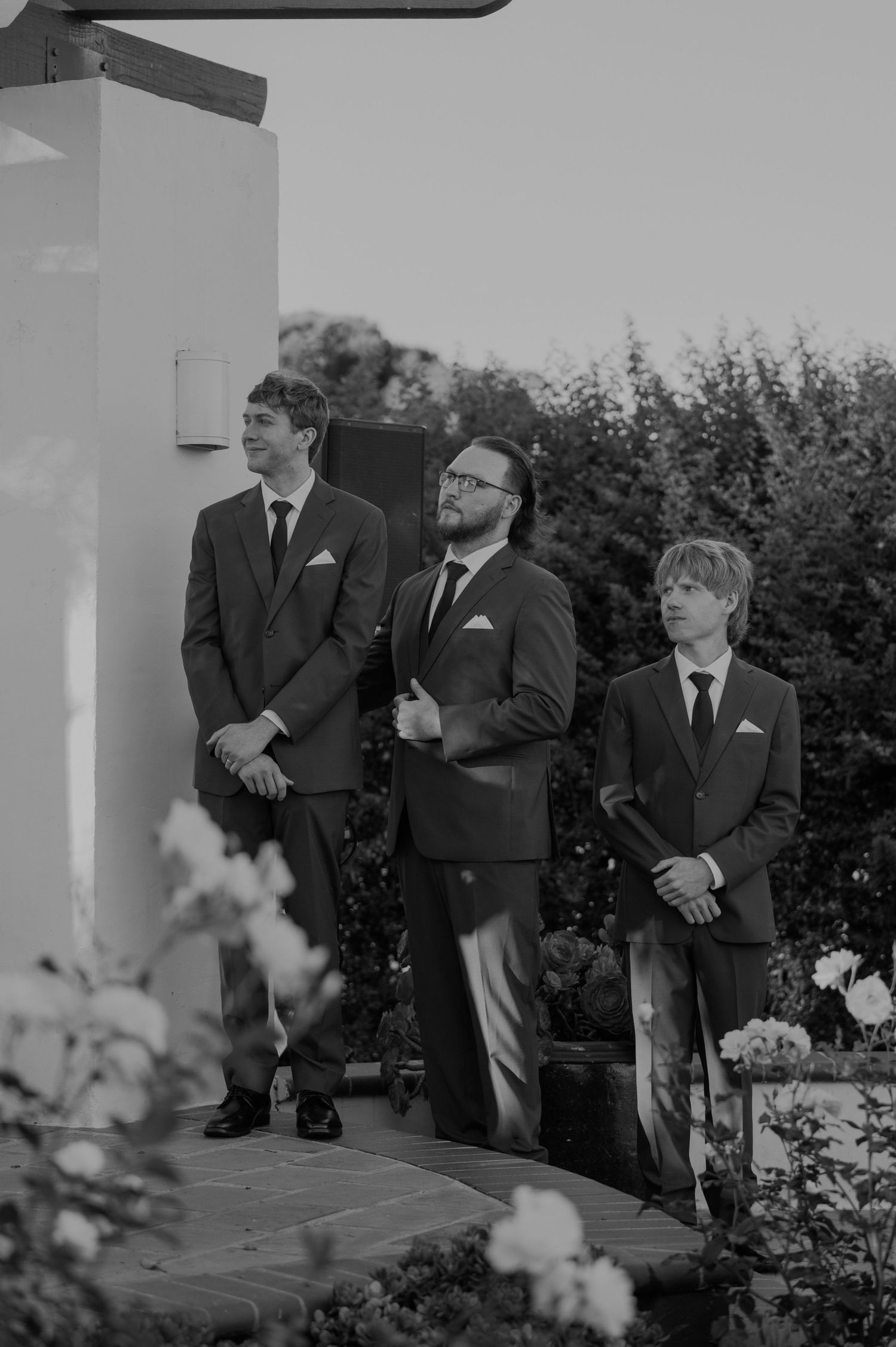 Three groomsmen in dark suits stand at outdoor wedding ceremony in black and white photo.