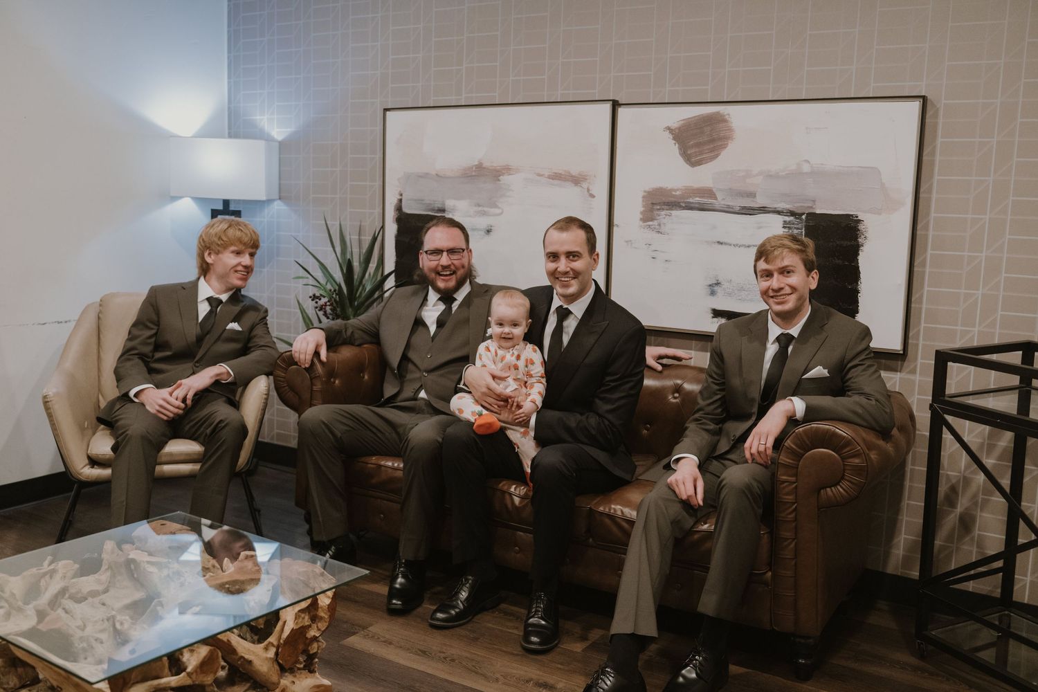Five men in matching brown suits pose for a photo in an elegant hotel lobby, with one holding a baby.