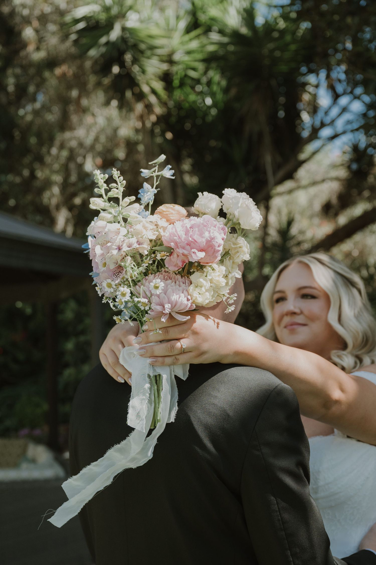 Bride in white top holds a pink and white wedding bouquet with ribbon streamers outdoors among lush greenery.