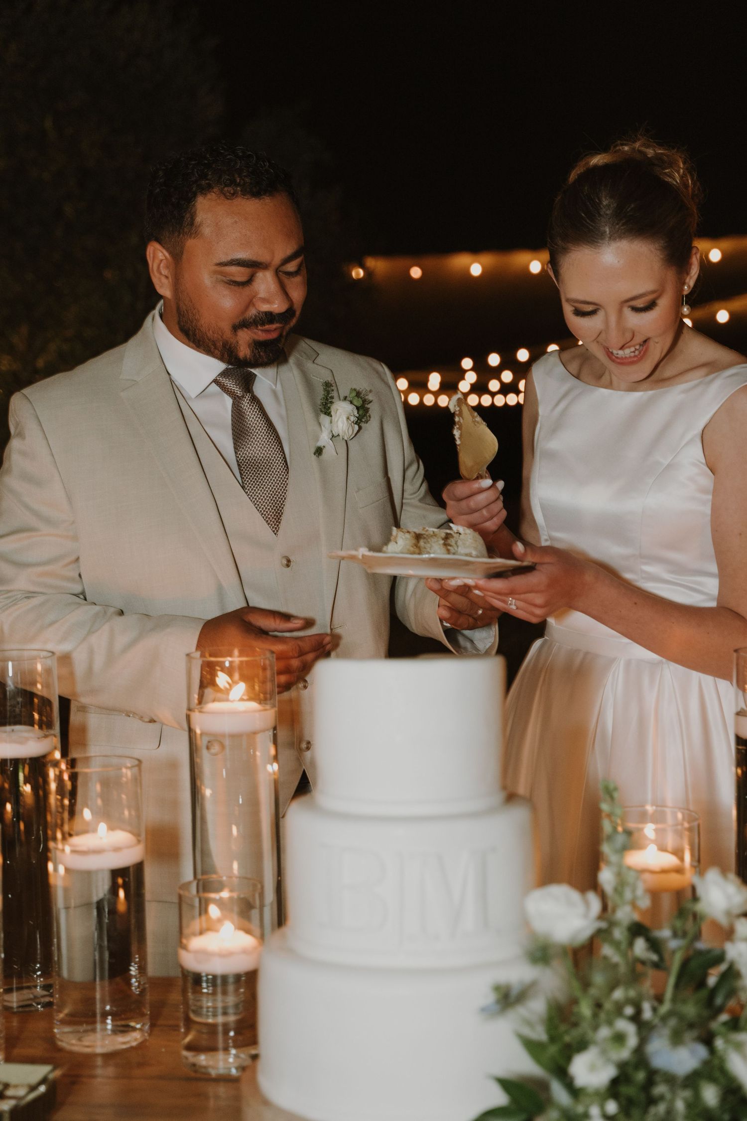 Newlyweds serving wedding cake slices beside candlelit three-tier white cake at evening reception.