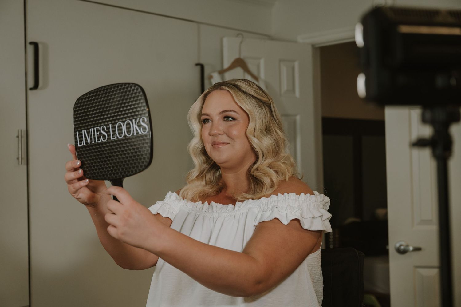 Woman in white off-shoulder top holding a Lives Looks branded mirror in front of a camera setup.