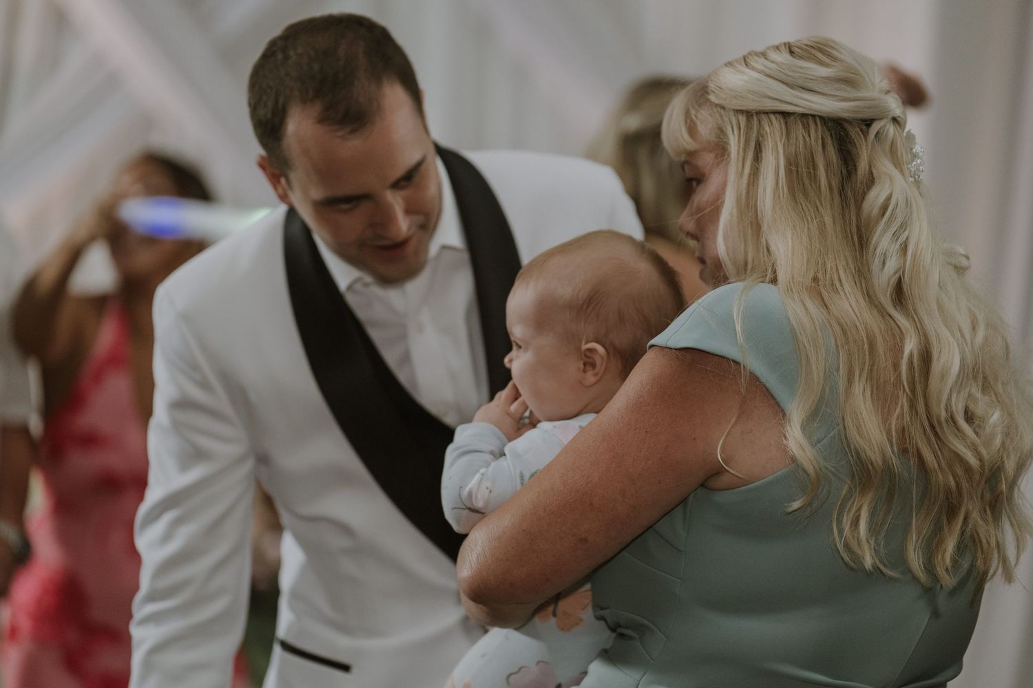 Groom in white tuxedo jacket admires a baby held by a blonde wedding guest during the reception celebration.
