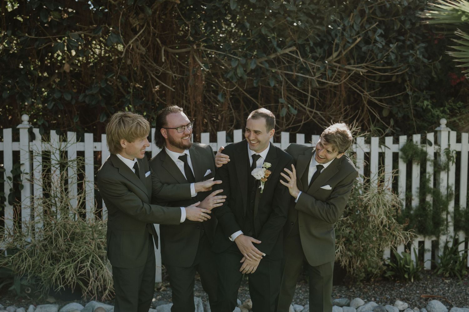 Four groomsmen in dark suits laughing and joking together outdoors near white picket fence.