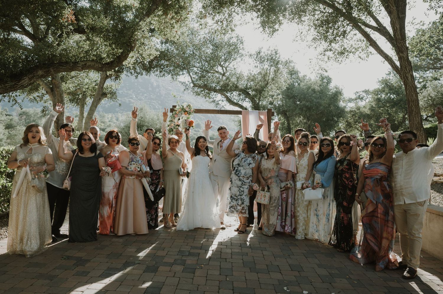 Joyful wedding party and guests celebrate outdoors with raised hands under shaded trees.