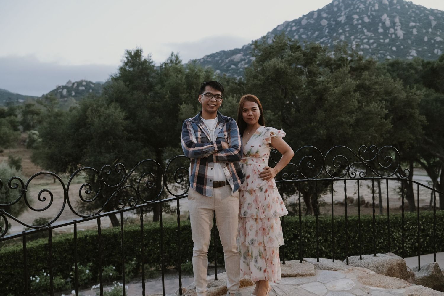 Couple posing together on a stone terrace with ornate iron railing and mountain views behind them.