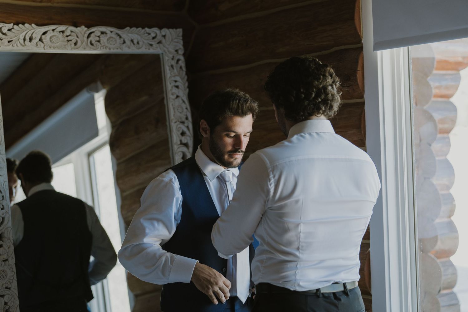 Groom getting dressed with groomsman help, adjusting vest and tie before wedding ceremony indoors.