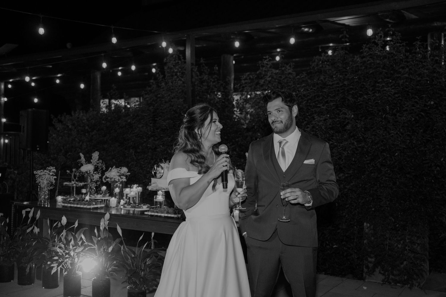 Bride and groom sharing a toast speech under string lights at an elegant outdoor wedding reception.