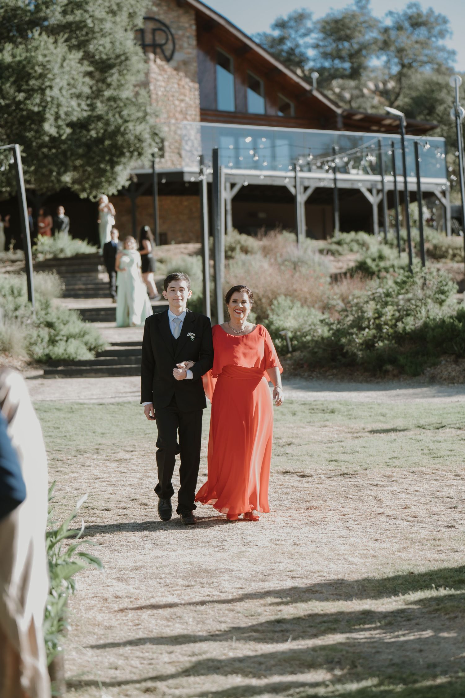 Young boy in black suit escorts woman in red gown at outdoor wedding ceremony.